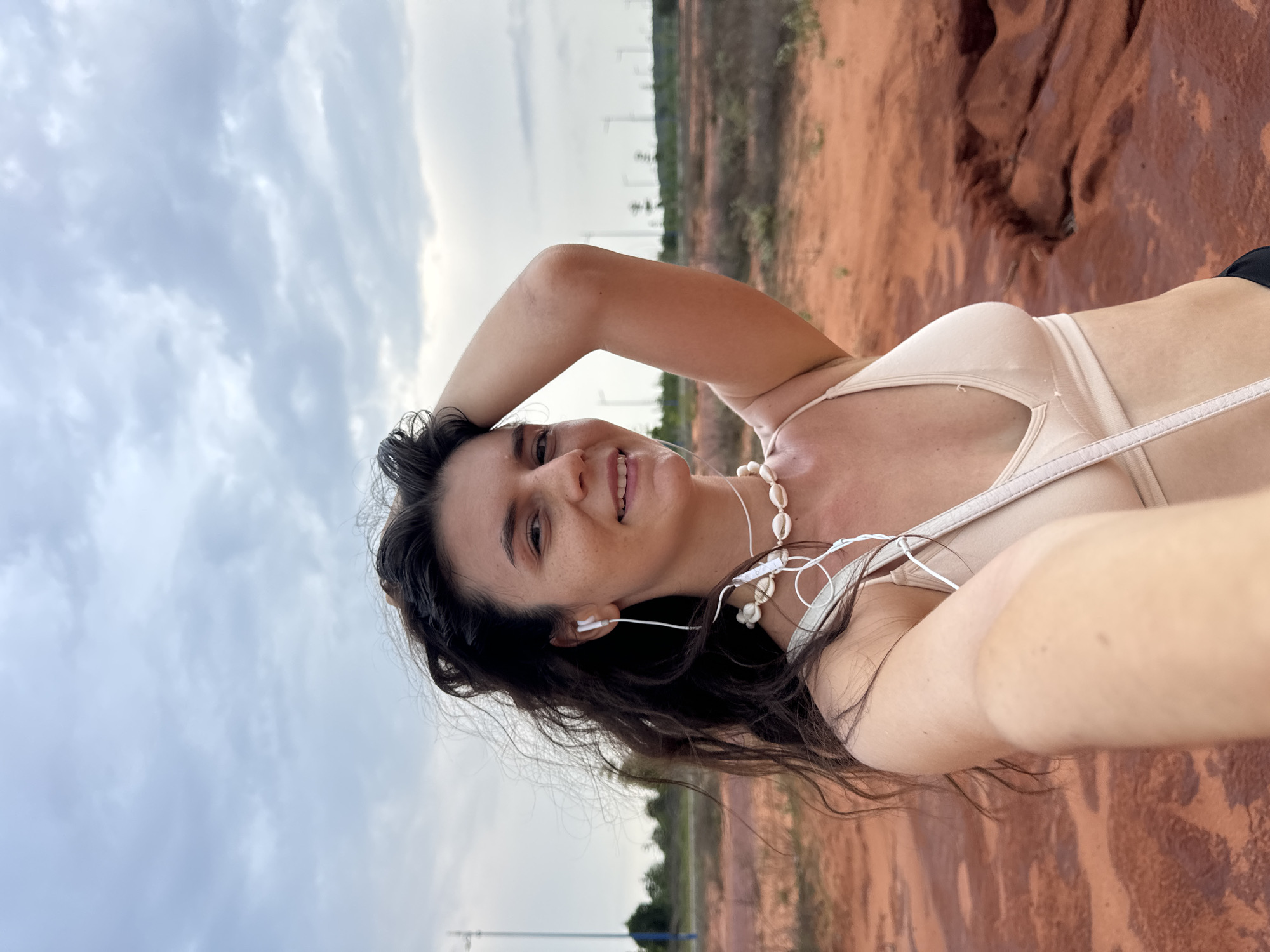 A young woman with long, dark, wavy hair is taking a selfie outdoors. She is wearing a light-colored bikini top and a shell necklace. Her left arm is raised behind her head, and she is smiling at the camera. In the background, there is an expanse of reddish-orange dirt and rocks, with a cloudy sky overhead. In the distance, several tall, slender structures, resembling wind turbines, can be seen against the horizon, along with sparse vegetation and what appears to be a road or pathway. The overall impression is a somewhat arid or undeveloped landscape.