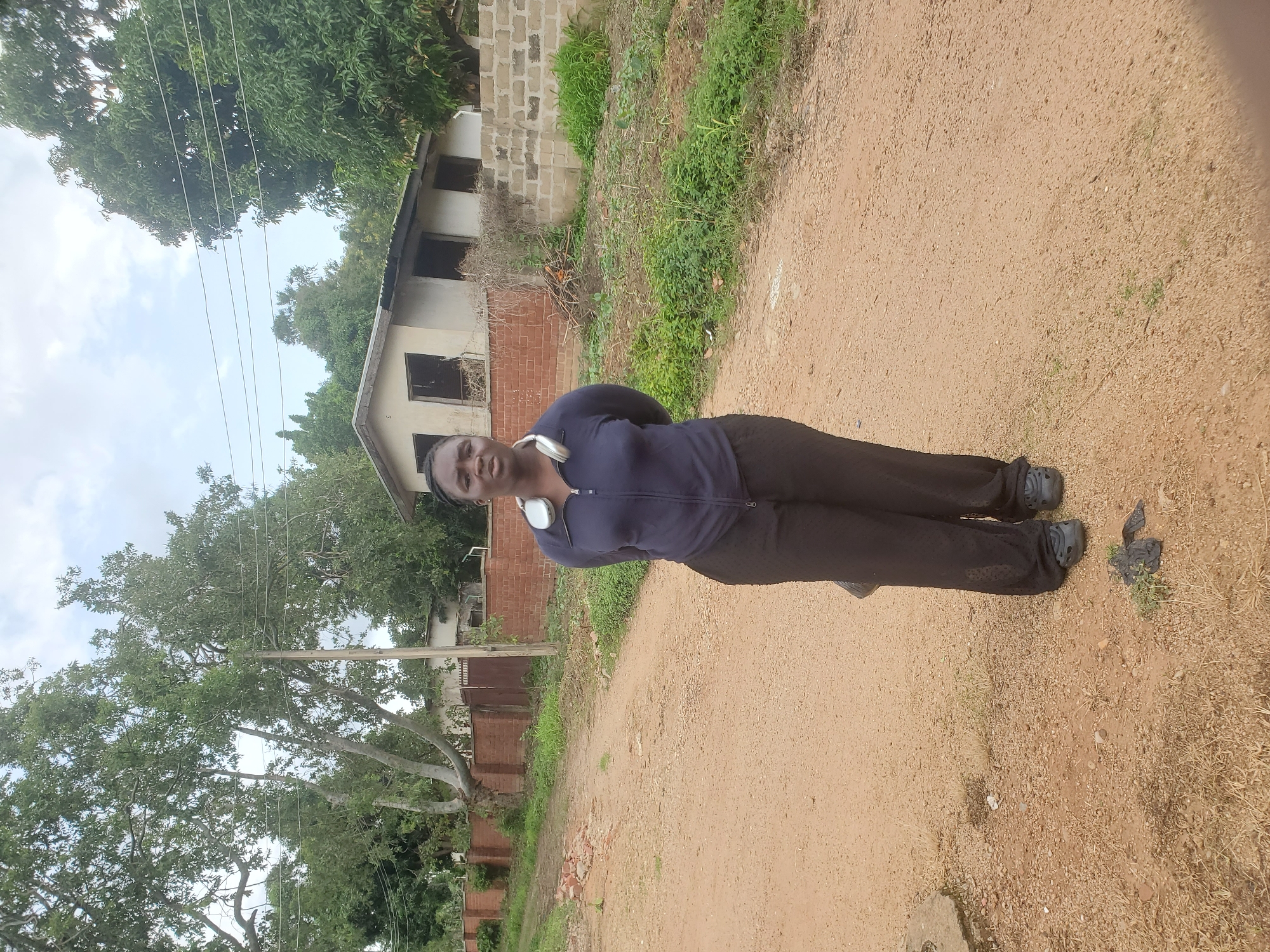 A full-body shot captures a Black woman standing on a dirt road in Jos, Nigeria. She is wearing a dark blue zipped jacket over a dark top and loose black pants. Silver headphones are around her neck. Her hands are clasped behind her back, and she is looking towards the camera with a neutral expression.
The background shows a rural or semi-rural setting. A weathered brick wall runs along the left side of the road, with trees and foliage behind it. Further back, a light-colored, single-story building with several windows is visible, surrounded by trees and shrubs. The road itself is unpaved, with scattered stones and dry grass. The sky is overcast with some visible clouds.