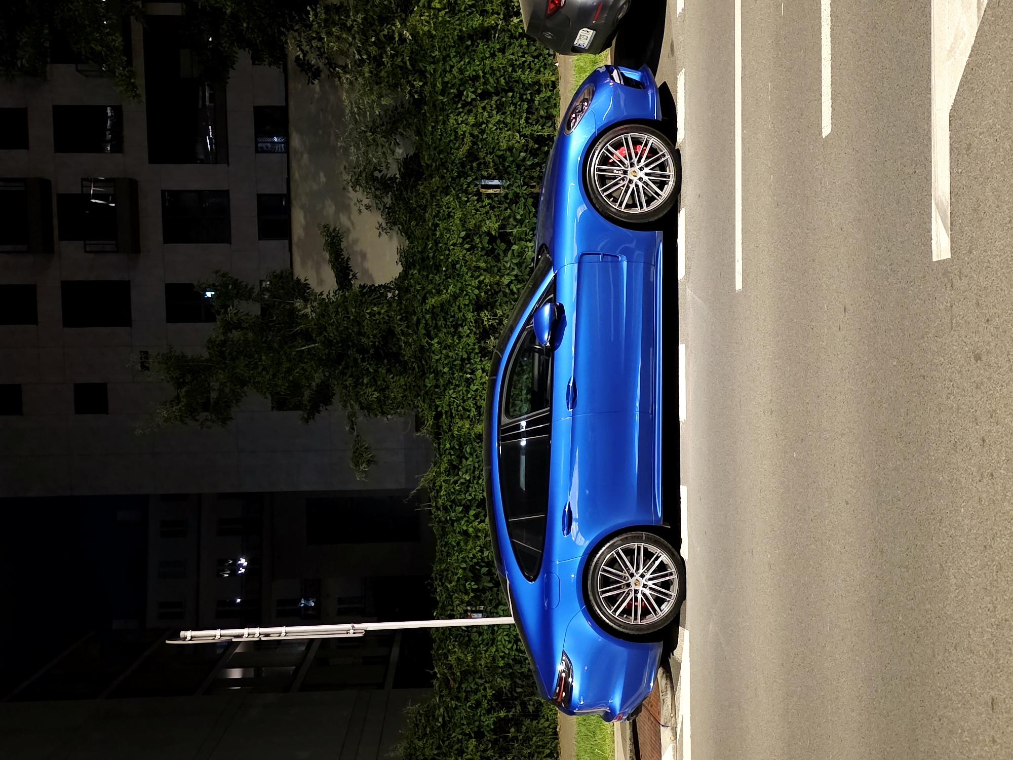 The image shows a blue Porsche Panamera parked on the side of a street at night in Warsaw, Poland. The car is positioned along a curb with a pedestrian crossing visible. In the background, there is a building with multiple windows and balconies, partially obscured by dense green foliage. The street is paved and appears to be a road in a residential or commercial area. No people are visible in the frame.