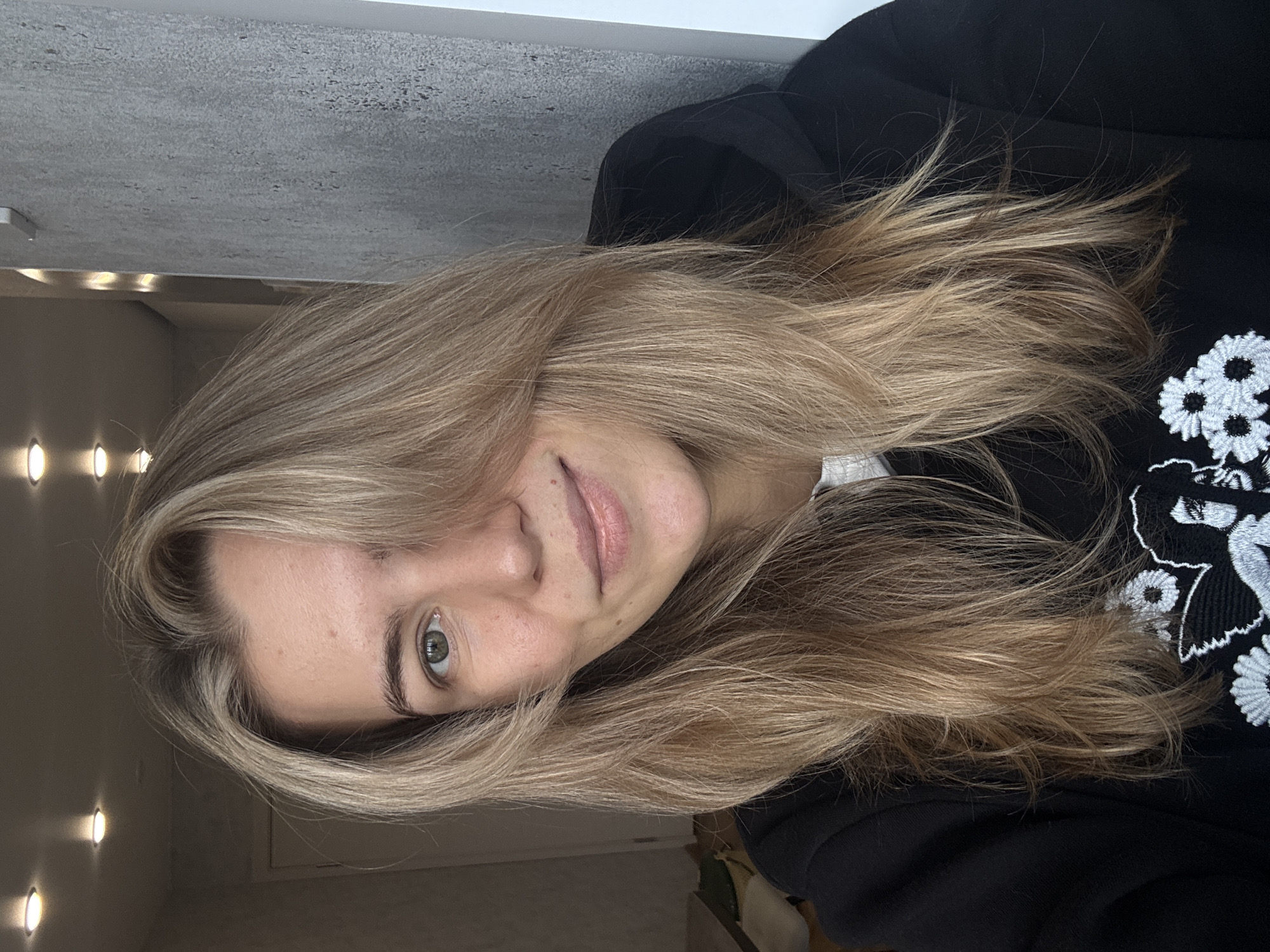 A woman with long, light brown hair is posing for a photo. She is wearing a black hoodie with white flower designs. The background is blurry but appears to be a room with a gray wall and a string of lights. The location is unknown.