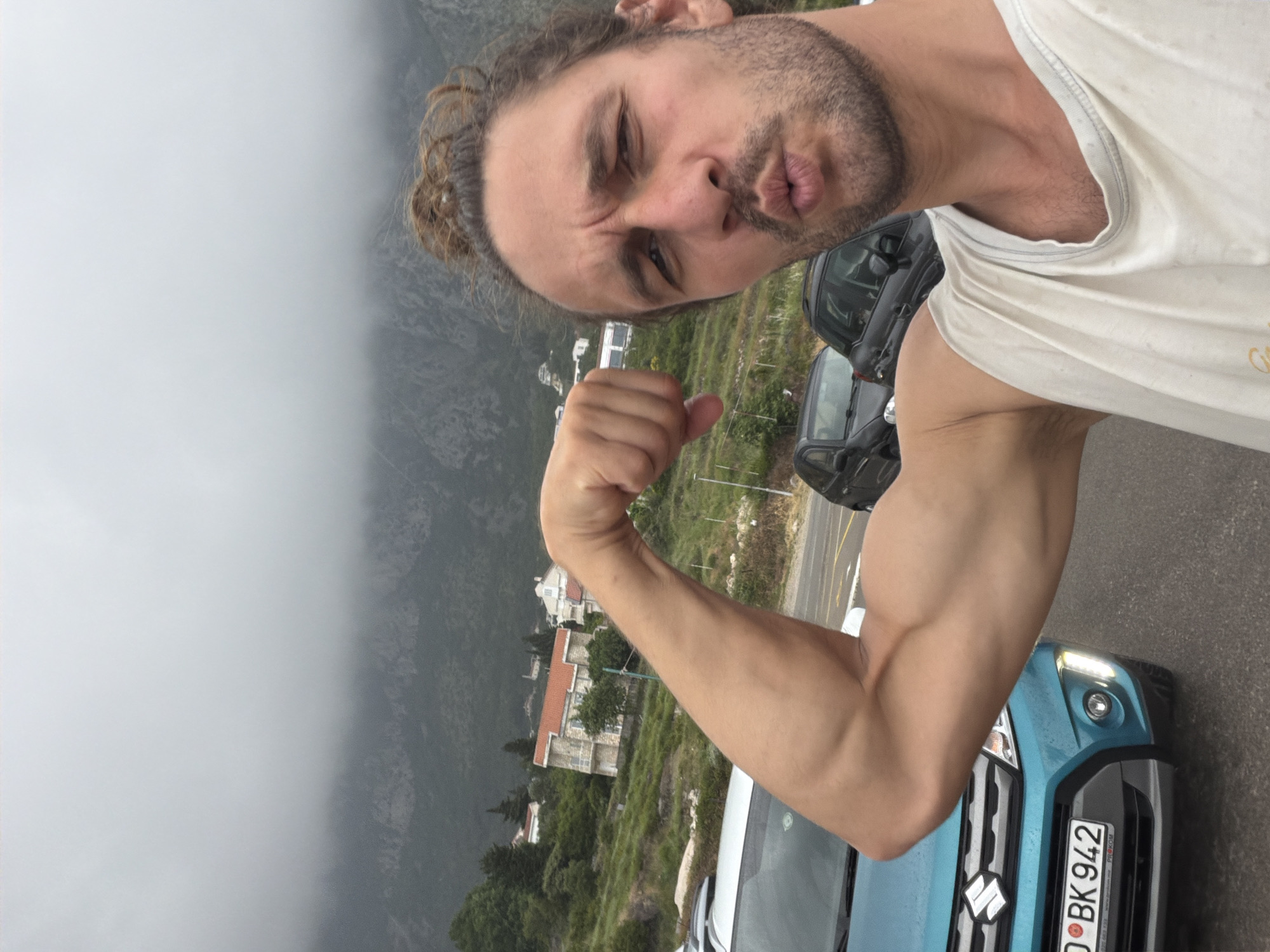 A man with light brown, curly hair tied back in a bun, and a beard, poses for a selfie, puckering his lips and squinting his eyes. He is wearing a white tank top and flexing his right bicep towards the camera, showcasing his muscular arm.
The setting is outdoors, in what appears to be a mountainous or hilly region, specifically Sveti Stefan, Montenegro, as indicated. The weather is overcast and foggy, with low-lying clouds or mist partially obscuring the distant mountains and sky, creating a muted color palette. Several buildings with distinctive red-tiled roofs are visible on a hillside in the mid-ground, surrounded by green vegetation.
In the foreground, a light blue (teal) Suzuki car is partially visible, with its front grille, headlights, and emblem clearly seen. Its license plate displays the text "0 BK 942" and "PREKOM". Another dark-colored car is partially visible further back on what appears to be a road or parking area. The man's playful expression and flexing pose suggest a lighthearted and confident mood.