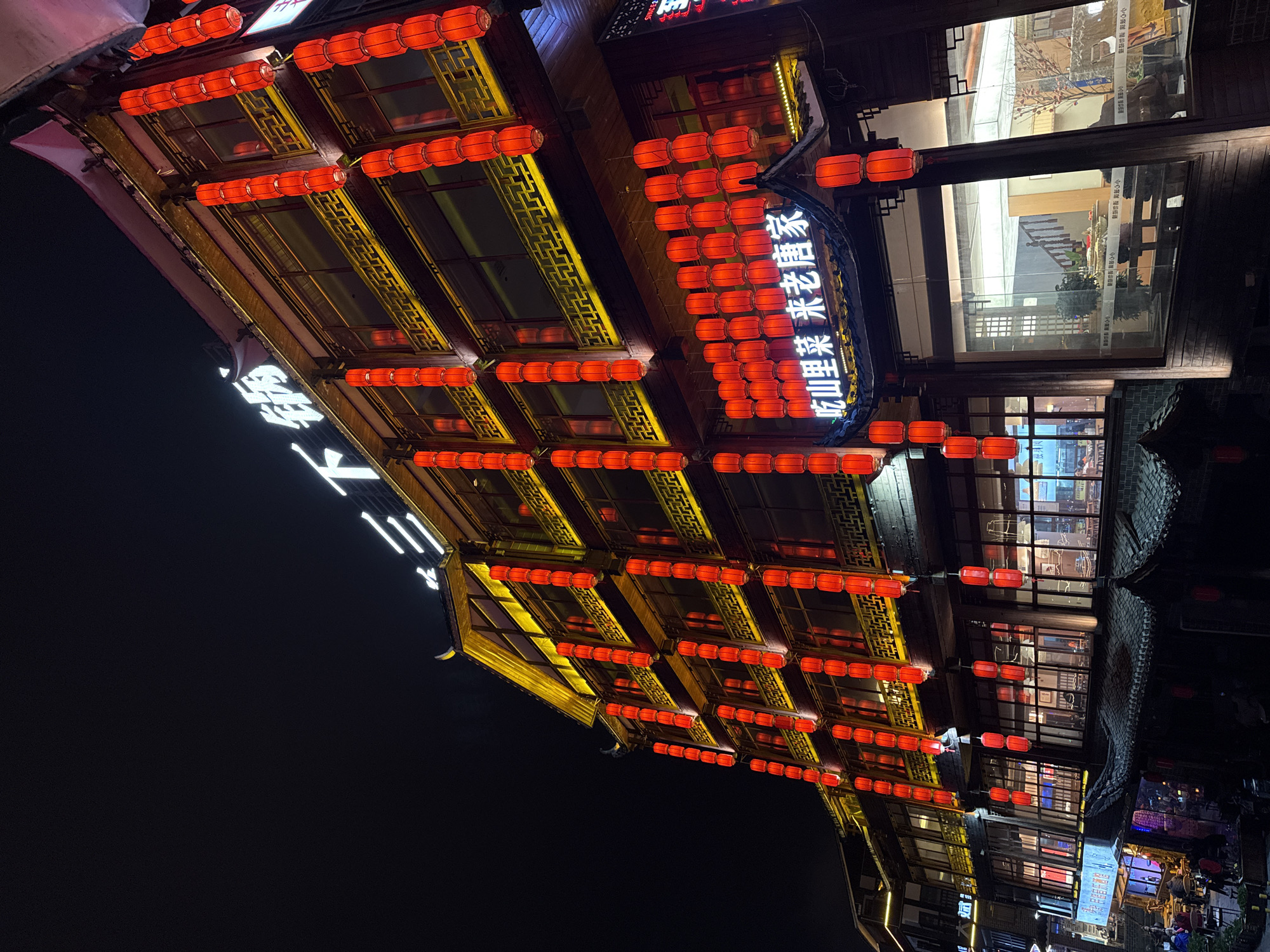 The image shows a building at night in Wulingyuan, China, adorned with rows of traditional red lanterns. The building's architecture features ornate, yellow latticework. A sign in Chinese characters hangs from the building and another vertical sign with illuminated characters appears on the left side. The background displays other structures and lights, suggesting a populated area.