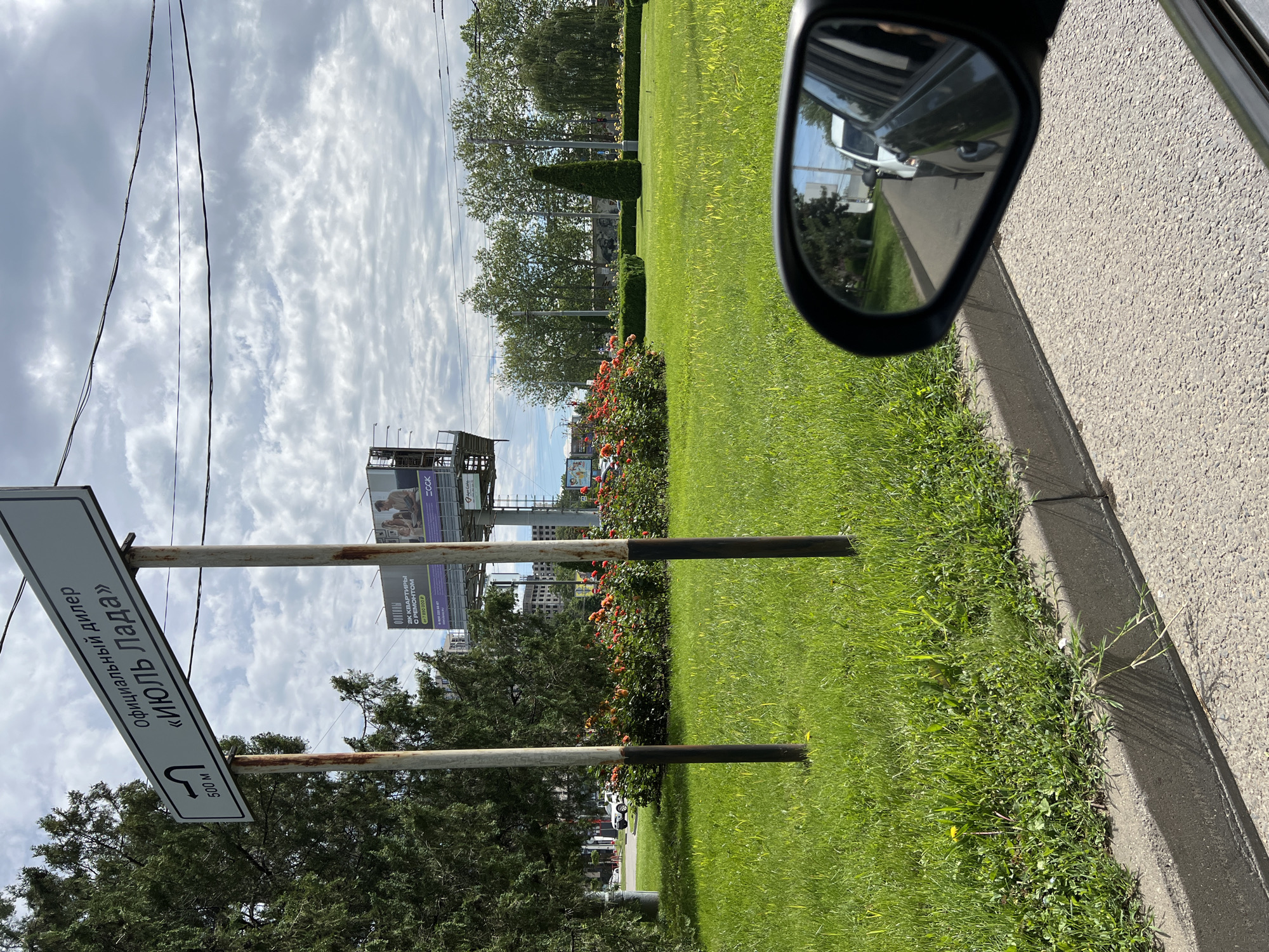 The image depicts a roadside scene in Krasnodar, Russia, captured from the perspective of a vehicle, looking out the passenger side window. The time of day appears to be daytime, with a bright, cloudy sky overhead.
In the foreground, a gray asphalt road shoulder and a concrete curb are visible on the right. A car's side mirror occupies the upper right, reflecting a white vehicle behind the camera and part of the street. To the left of the curb, a wide expanse of vibrant green grass stretches across the frame, indicating a well-maintained roadside landscape. This grassy area features manicured hedges and several rose bushes with orange-red blooms. Various trees, some with light green foliage, are scattered throughout the midground and background, and thin power lines crisscross the sky.
A prominent street sign, attached to two rusty metal poles, is positioned in the midground. The sign, written in Cyrillic, states "Официальный дилер «Июль Лада»" (Official dealer "July Lada") and includes a left-curved arrow along with the text "500 м" (500 meters), indicating the direction and distance to the dealership. Behind the sign and landscaping, two billboards are visible. One partially displays the text "ССК" and an image. The other advertisement reads "2К КВАРТИРЫ С РЕМОНТОМ" (2-room apartments with renovation) and "от 7 890 000" (from 7,890,000), followed by phone numbers. The scene portrays a typical urban or suburban environment with commercial advertising and clear navigational signage.