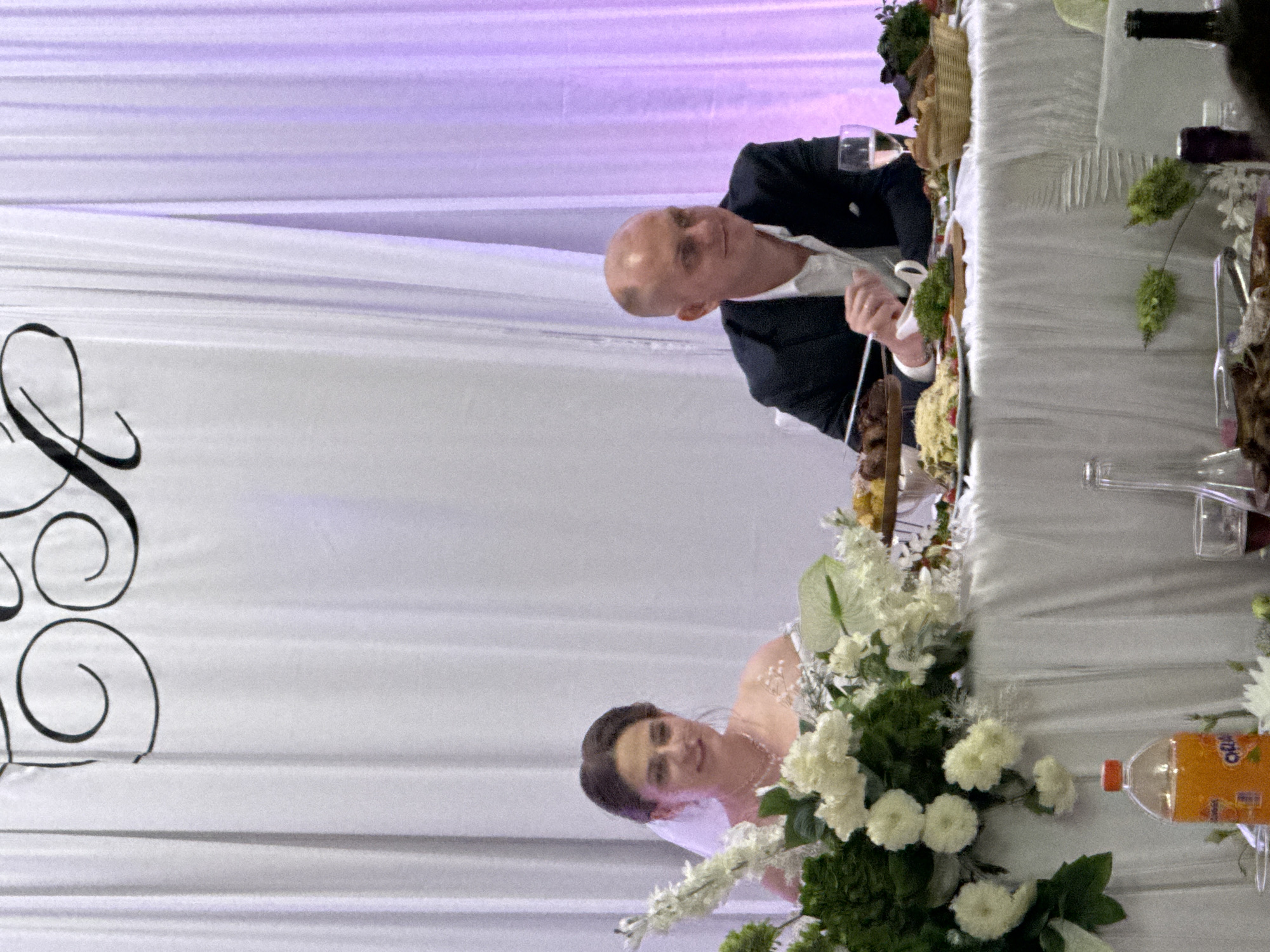 A bride and groom are seated at a draped table, likely at a wedding reception. The bride, on the left, wears a white wedding dress and veil, with a necklace. She is smiling and looking towards the viewer. The groom, on the right, wears a dark suit jacket over a white shirt and is looking to his right. The table is adorned with white flowers and green foliage on the left, and a variety of food dishes, a basket of bread, and a glass of white wine on the right. In the background, white and purple fabric drapes create a soft, illuminated backdrop. A bottle of orange soda is visible on the left side of the table. The overall setting suggests a formal celebratory event. The location is identified as Ust-Labinsk, Russia.