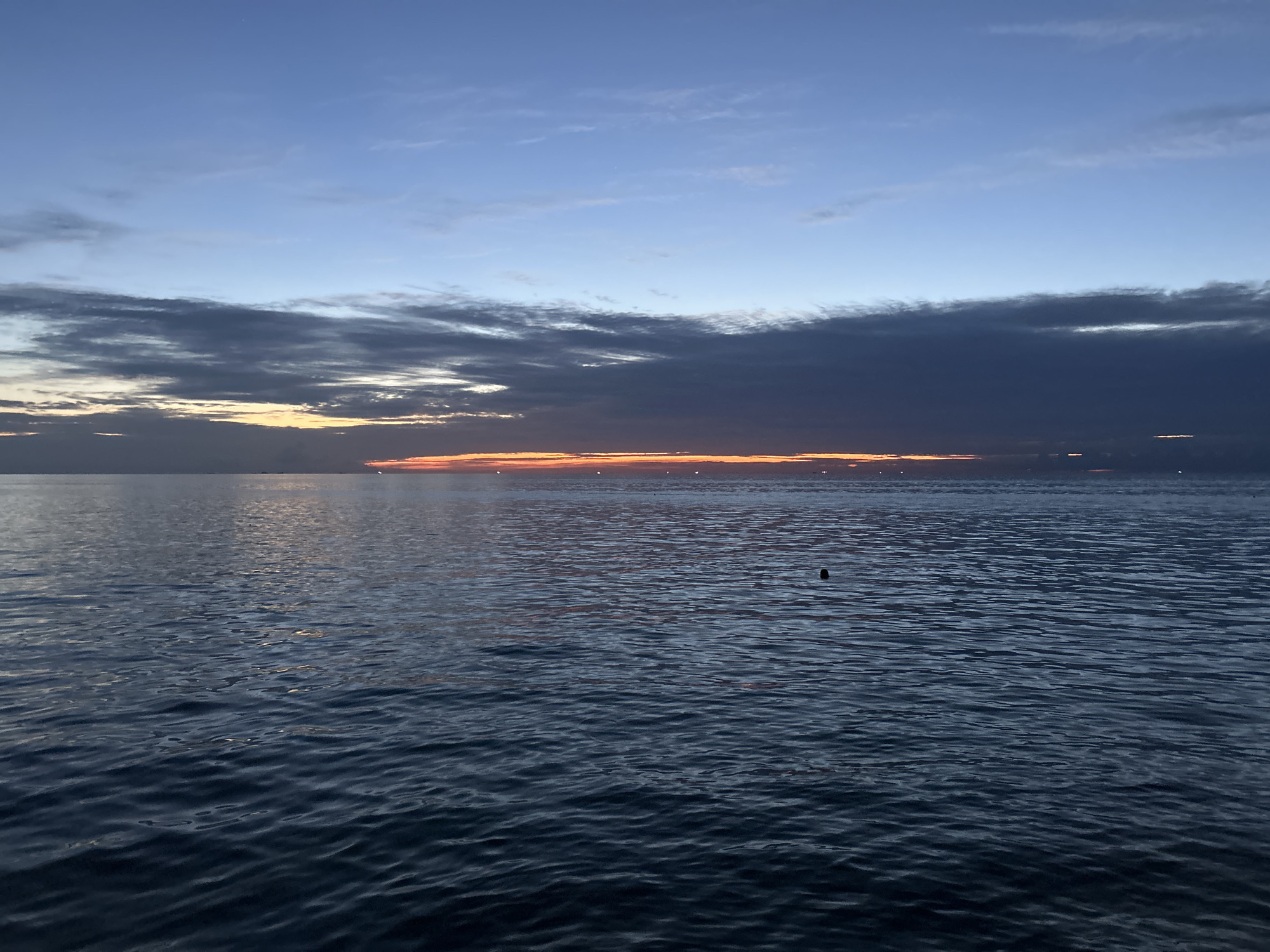 The image depicts a serene seascape at twilight, likely taken from the shore or a vessel near Ko Phangan, Thailand. The sky transitions from a clear, pale blue at the top to darker, dramatic clouds tinged with orange and yellow along the horizon, indicating the setting or rising sun. The sea is calm with gentle ripples reflecting the soft colors of the sky. A small, dark, spherical object, possibly a buoy or marker, is visible in the middle right of the frame, floating on the water. The overall atmosphere is tranquil and peaceful, characteristic of a quiet evening or early morning by the sea.