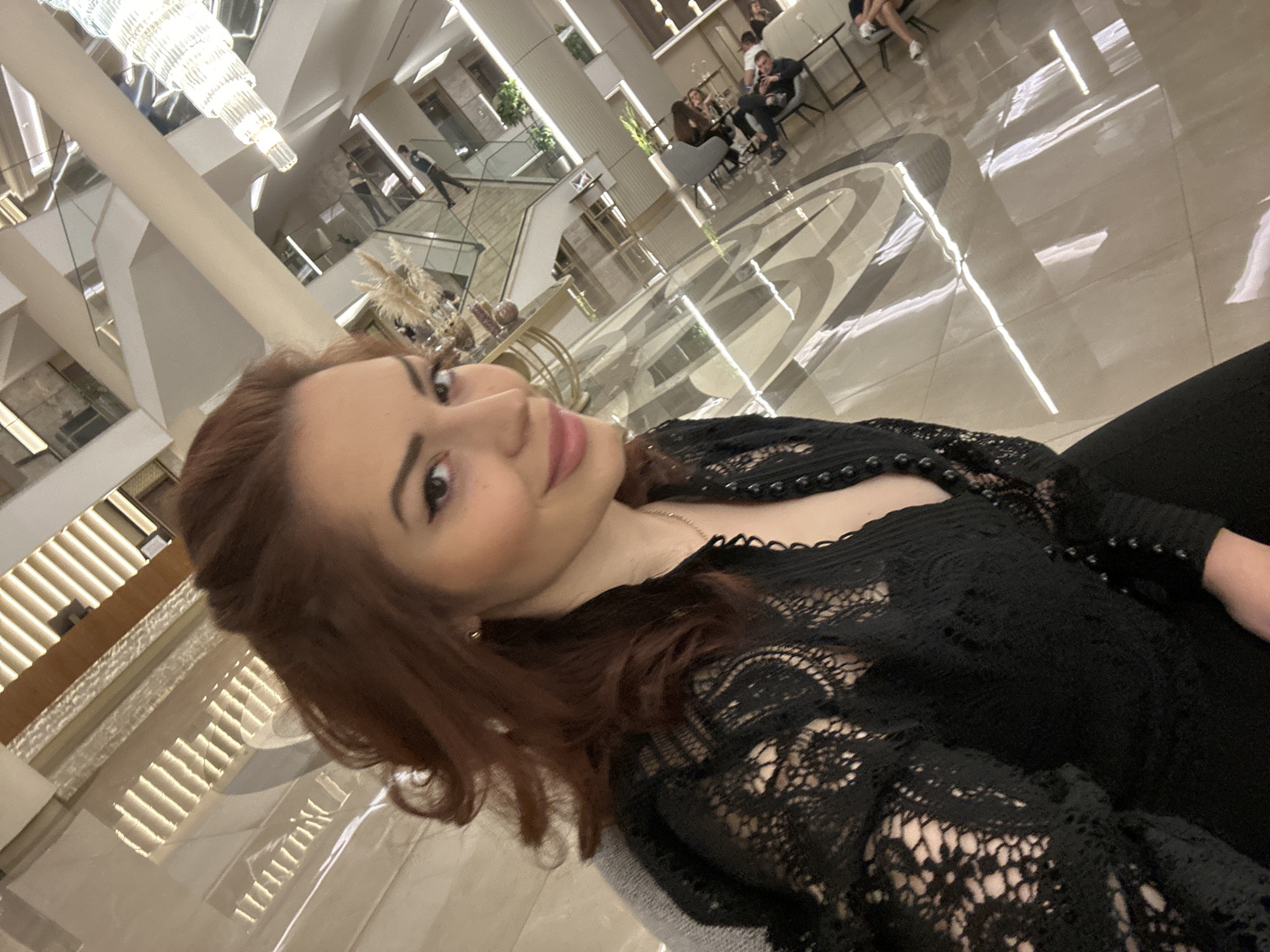 A young woman with reddish-brown hair and dark eyes, wearing a black lace top with pearl embellishments, is captured in a close-up selfie from a low angle, slightly tilted. She has light makeup and is looking towards the viewer with a subtle smile. A thin gold necklace is visible around her neck.
The background reveals a spacious, modern, and upscale interior, likely a hotel lobby or a grand public building in Kazanlak, Bulgaria. The setting features high ceilings and a polished, light-colored floor with dark geometric patterns, reflecting the bright overhead lighting. A large, multi-tiered chandelier hangs prominently from the ceiling. The space extends over multiple levels, connected by staircases with glass railings.
Several people are visible in the mid-ground and background. Some are seated at modern tables and chairs in a lounge area, while others appear to be walking up or down the stairs. Decorative plants or dried arrangements add to the elegant decor. The overall lighting is bright and artificial, creating a well-lit and inviting ambiance. There is no visible text in the image.