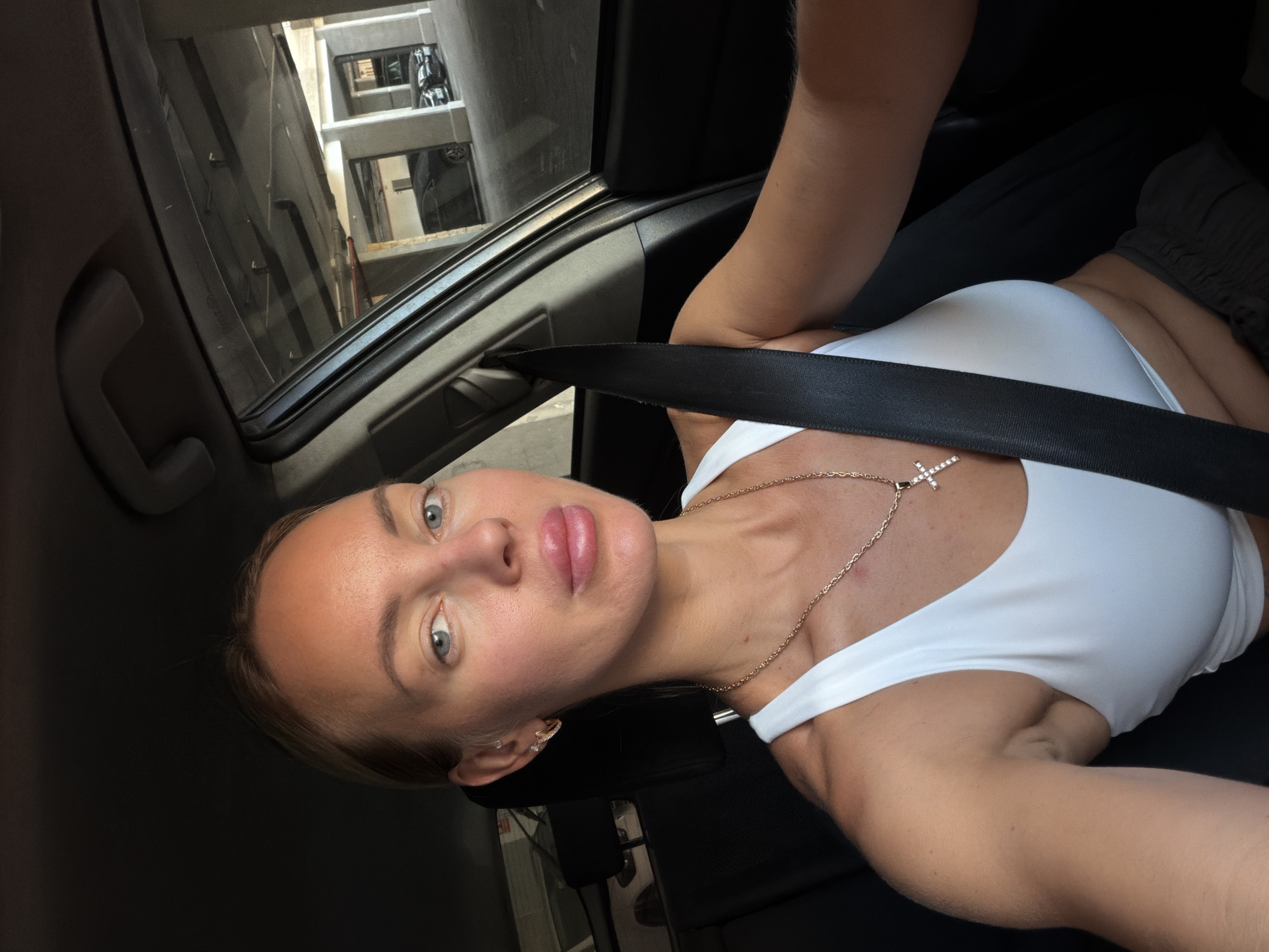 The image shows a woman inside a car, with a seatbelt fastened. She is looking at the camera and appears to be in a parking structure. The background shows concrete pillars and other vehicles, suggesting an urban environment. The lighting is bright, indicating it is daytime.