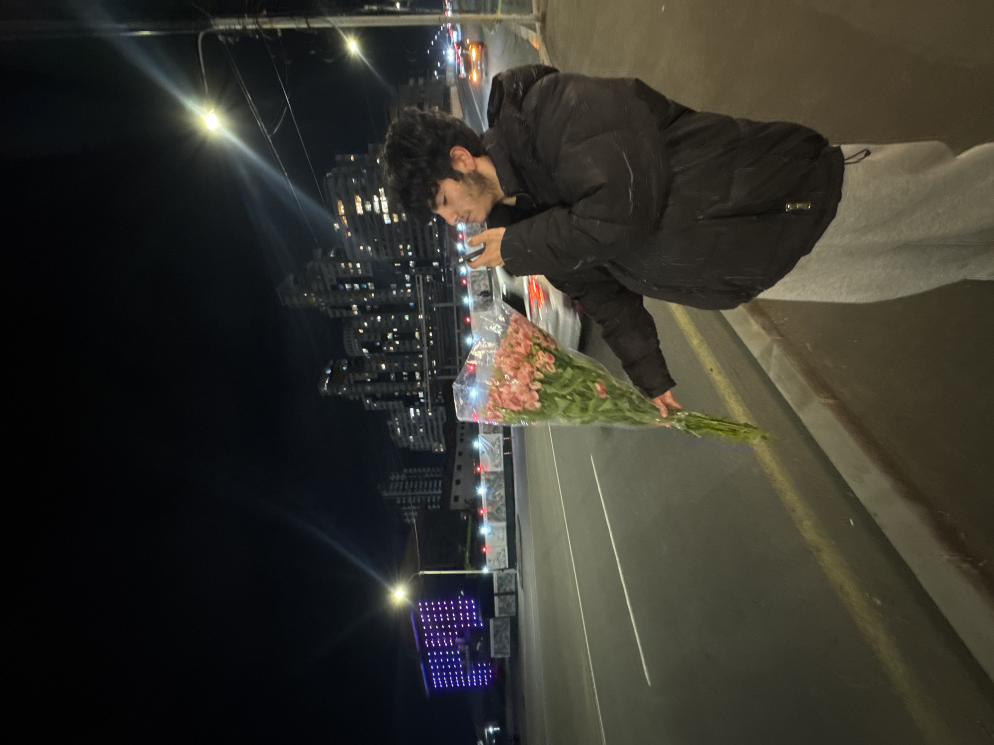 A man stands on a sidewalk next to a street in Yerevan, Armenia at night. He holds a bouquet of pink flowers in plastic wrapping, and appears to be looking at his cell phone. He is wearing a black jacket and grey sweatpants. Behind him, a city skyline of tall buildings is visible, illuminated by lights. Streetlights line the road, and a small lit up purple art installation is also visible. Cars are driving on the street.