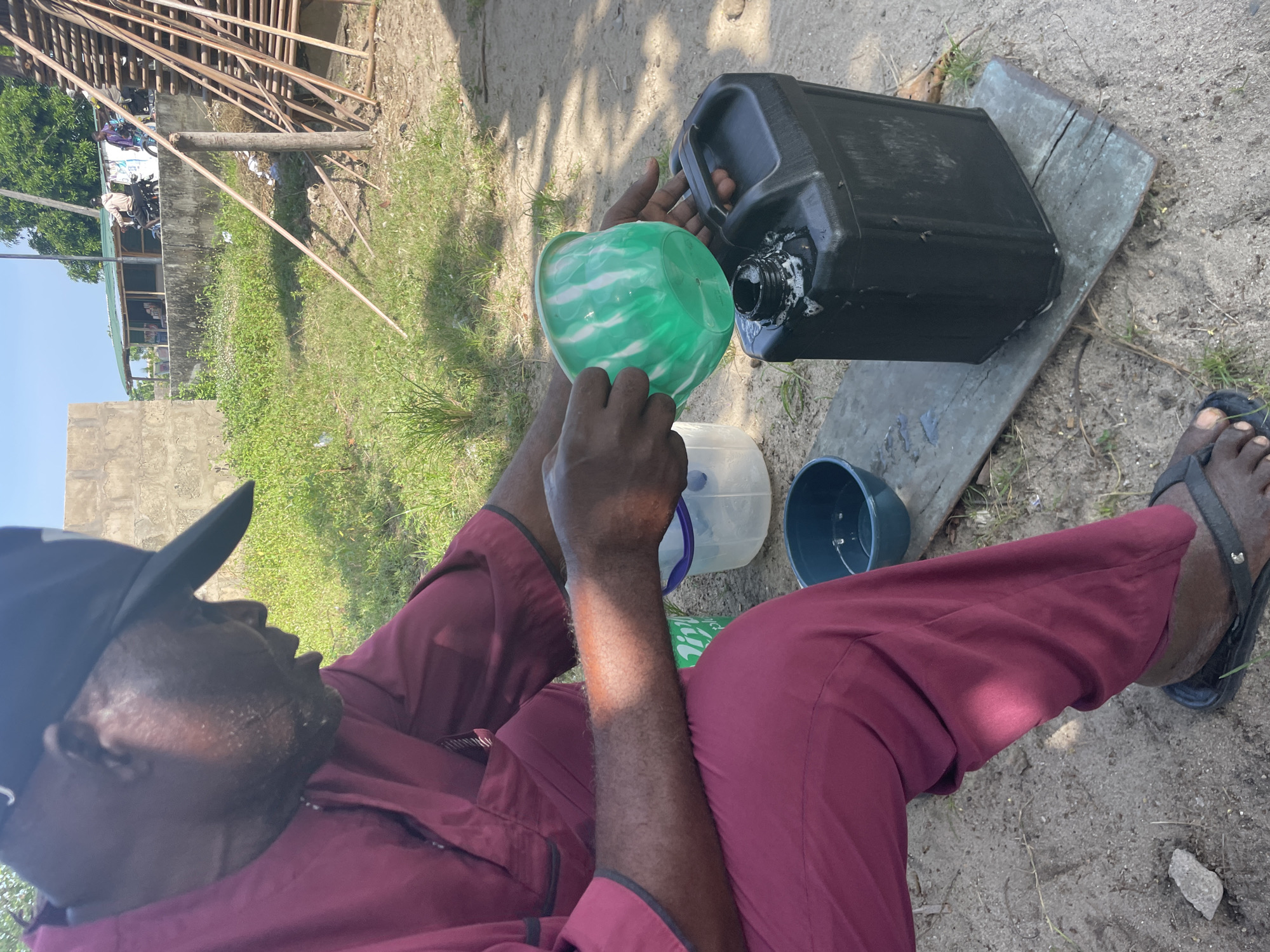 This image, set in Badagry, Nigeria, depicts an adult male engaged in an outdoor task under bright daylight. The man, wearing a dark baseball cap, a maroon long-sleeved shirt, and black flip-flops, is seated on the ground. The ground is a mix of loose dirt, small rocks, and patches of green grass.
The main activity shows him carefully pouring liquid from a large, black plastic jerrycan into a smaller, green and white marbled plastic bucket which he holds. The jerrycan rests on a wooden plank and has noticeable white, foamy residue around its spout, suggesting the contents could be soapy water or a frothy liquid. A smaller, translucent plastic container is partially visible behind his hands, and a dark blue plastic bowl sits on the ground near his feet.
The background reveals a somewhat rustic or informal outdoor setting. On the left, there is a concrete wall, and further back, a building with a green roof is visible, along with green vegetation and what appears to be laundry hanging. Above, a structure of wooden beams, possibly scaffolding or part of a building frame, is prominent. Strong sunlight casts clear shadows, indicating it is likely mid-morning or afternoon. The scene portrays an everyday task in a seemingly undeveloped or casual environment. No discernible text is visible on any objects.
