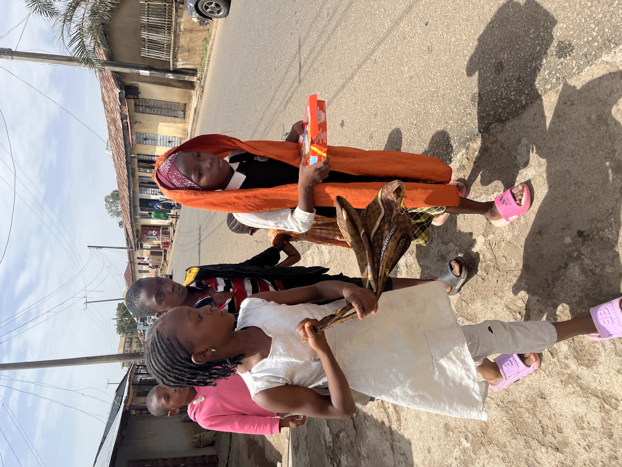 In a medium-shot, a group of African children stand on a paved street in Jos, Nigeria, bathed in bright sunlight. The lead child wears a white dress and grey trousers, lavender-colored slides and has braided hair. The child holds a scarf in their hands. Behind them, another child is partially visible in a pink shirt. Further back, a boy in a yellow and black jacket is seen, followed by another child. The child at the back has their head wrapped in orange fabric, wears a dark jacket and holds a red box. The residential buildings are visible in the background along the left side of the frame. A car is parked on the right side of the image. The scene is captured from a slightly elevated angle.