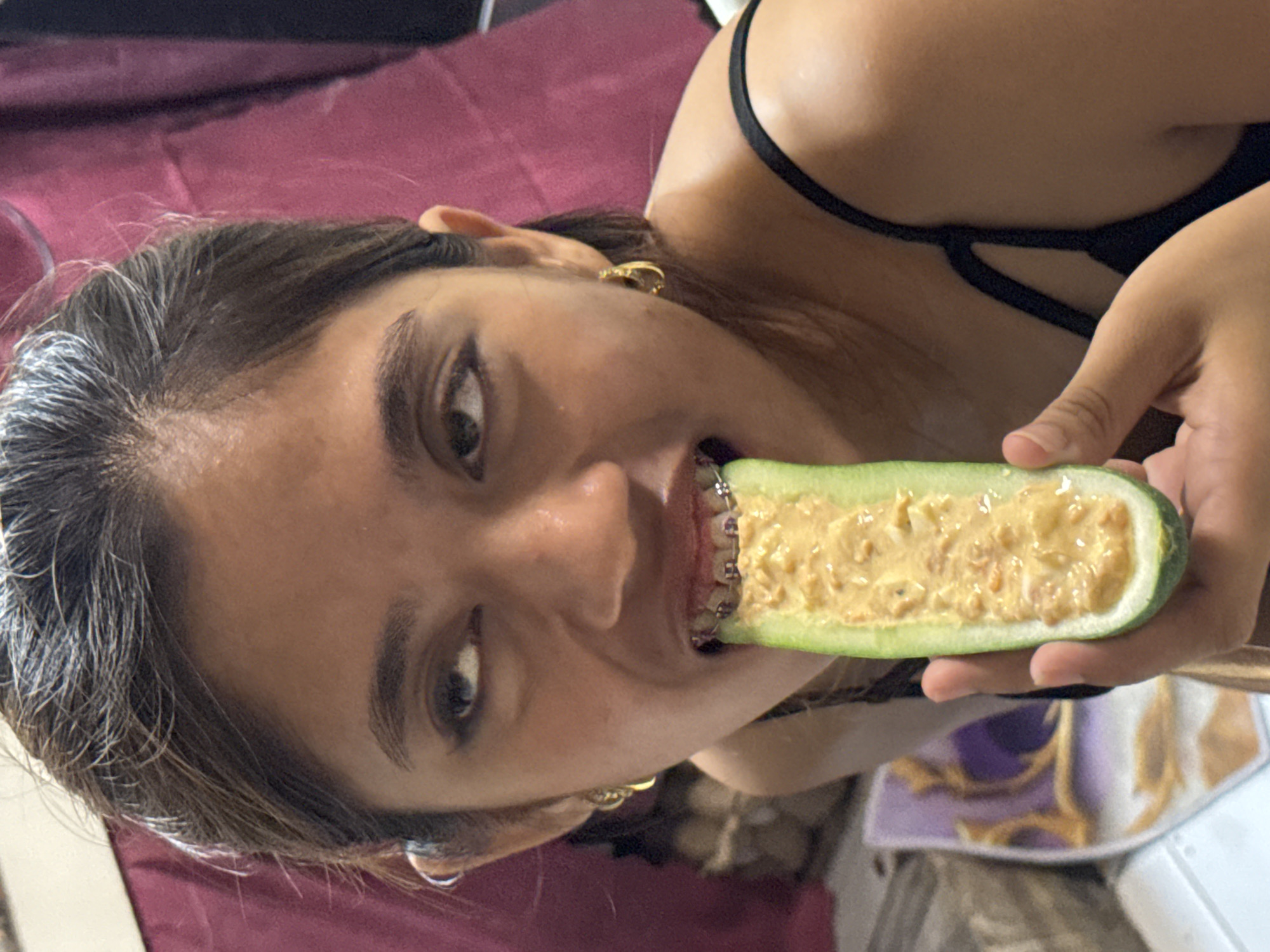 A young woman with dark hair pulled back and wearing gold hoop earrings, a black camisole, and dental braces, is shown from the chest up. She is holding a halved cucumber filled with a creamy, yellowish substance, with the cucumber pressed against her open mouth. Her eyes are looking upwards and to the left, with eyeliner visible. The background consists of a blurry maroon curtain and a sliver of a patterned object on a white surface. The scene appears to be indoors, possibly in a residential setting in Davao City, Philippines.