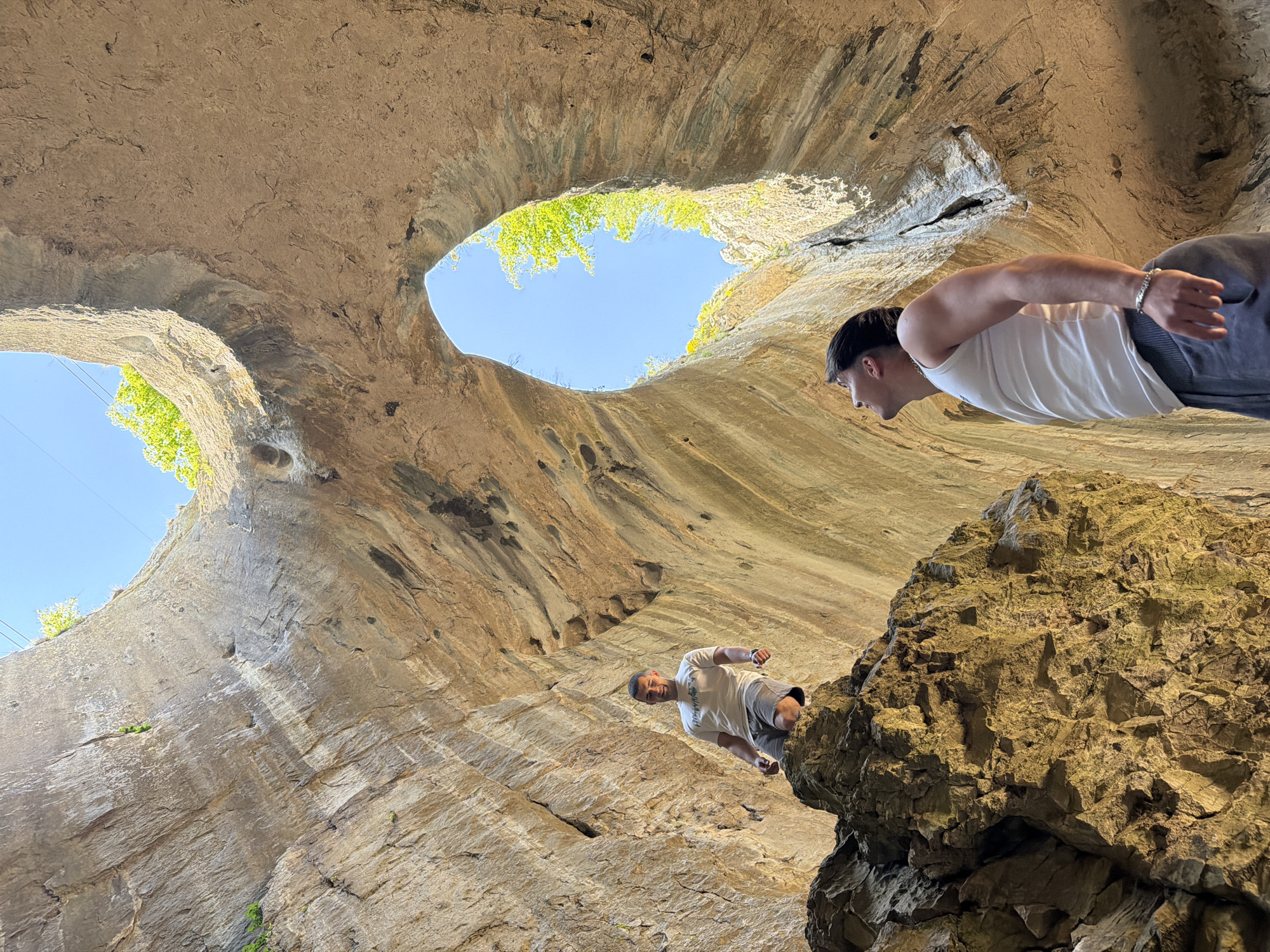 The photograph depicts a rock formation in Karlukovo, Bulgaria, with two large circular openings in the rock, allowing views of the sky and vegetation above. The rock walls are textured and appear layered. Two individuals are visible: one standing on a rocky outcrop, facing down, and another positioned higher and to the right, appearing to be looking into the opening of the rock formation. The scene suggests a natural rock cave or arch structure with openings allowing light and views of the outside.