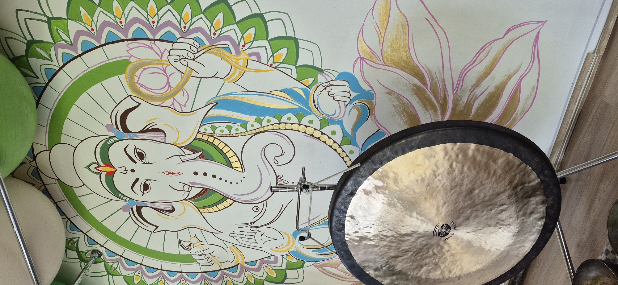 A medium shot depicts a gong positioned in front of a wall featuring a painting of Ganesha, set against a background with colorful patterns and floral motifs. The gong is the dominant element in the foreground, exhibiting a metallic sheen and a dark outer rim. Visible on its surface is a logo or inscription. The wall behind the gong serves as a backdrop, adorned with a colorful illustration of Ganesha, a Hindu deity, and additional artistic details. The environment suggests an indoor setting, potentially a studio, meditation space, or residence, though no specific location information is provided. The overall scene is suggestive of a creative or spiritual ambiance.