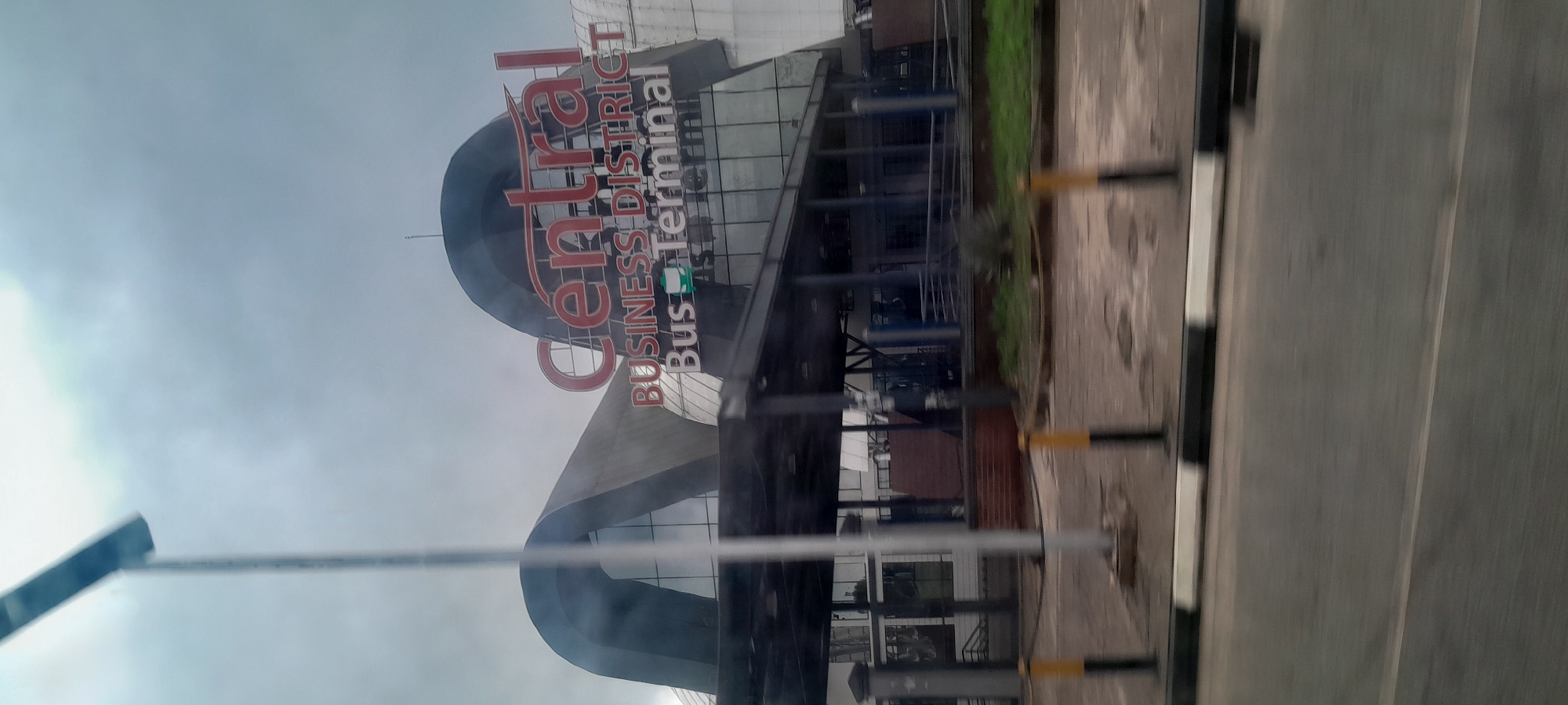 The image shows the Central Business District Bus Terminal in Abuja, Nigeria. The building has a modern architectural design with curved roofs and large glass panels. A prominent red sign reads "Central BUSINESS DISTRICT Bus Terminal". The sky is overcast with clouds. In the foreground, there is a road and a sidewalk with bollards. The scene appears to be captured from a moving vehicle, as indicated by motion blur.