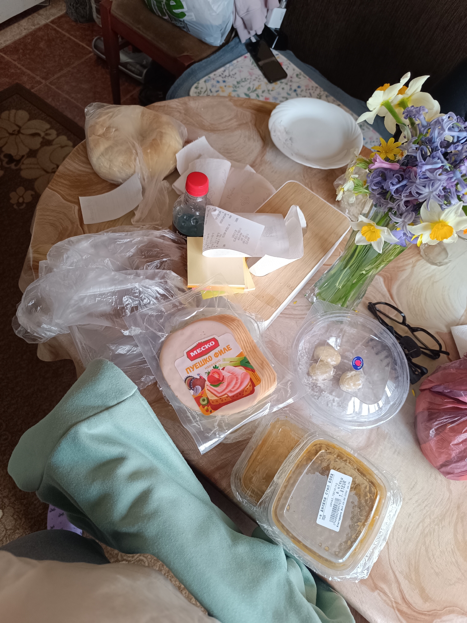 The image shows a cluttered tabletop with various items, suggesting a recent shopping trip or meal preparation. At the center is a package of "MECKO ПУЕШКО ФИЛЕ" (turkey fillet) and a container of "ДИЛЕМА СТИЛ ЕООД" hummus. Other items include a plastic bag containing bread, a small bottle with a red cap (possibly a condiment or dressing), receipts, cheese slices, plastic bags, a wooden cutting board, a white plate, a vase of flowers, a container with what appears to be small dumplings, and eyeglasses. A light green garment is partially visible on the left side. The items suggest a location in Bulgaria, specifically Pleven, based on the brand names and the label on the hummus. The overall impression is of a casual, domestic scene.