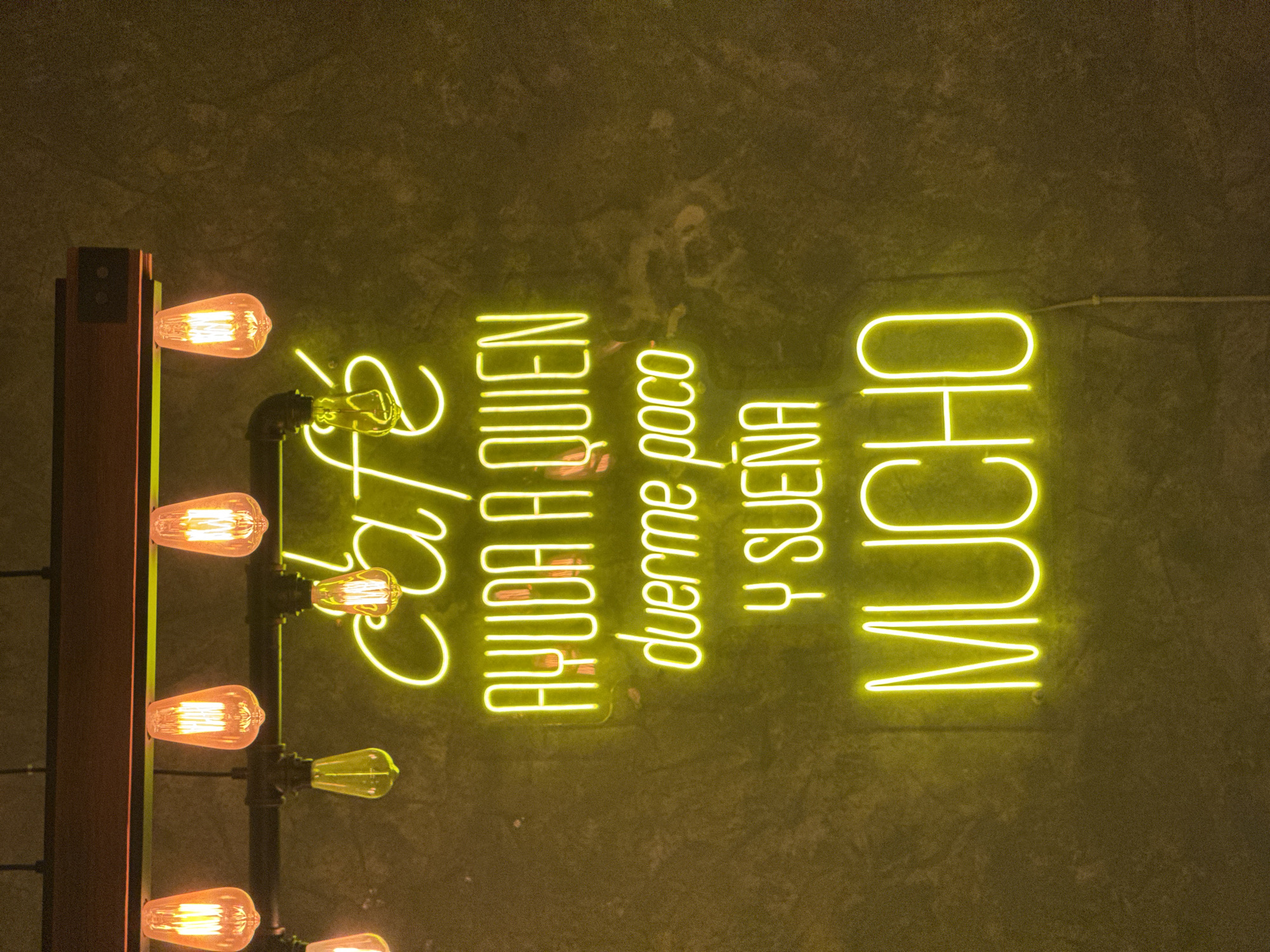 The image depicts a dimly lit interior scene, possibly in Puebla, Mexico, as indicated by the Spanish phrase "Café, ayuda a quien duerme poco y sueña mucho" (Coffee helps those who sleep little and dream much). This suggests a café or restaurant setting.
The scene features a wall with a concrete-like texture. On the wall, a bright yellow neon sign displays the Spanish phrase. To the left of the neon sign is a vertical structure, possibly a part of the wall decoration, with a wooden frame and a series of vintage-style light bulbs attached via metal pipes. The bulbs are glowing, casting a warm light.
There are no people visible in the image, and the primary subjects are the neon sign and the light fixture. The interaction between the objects is that the light fixture is illuminating the area and potentially drawing attention to the neon sign. The environment appears to be a modern or industrial-style interior with a focus on visual elements.