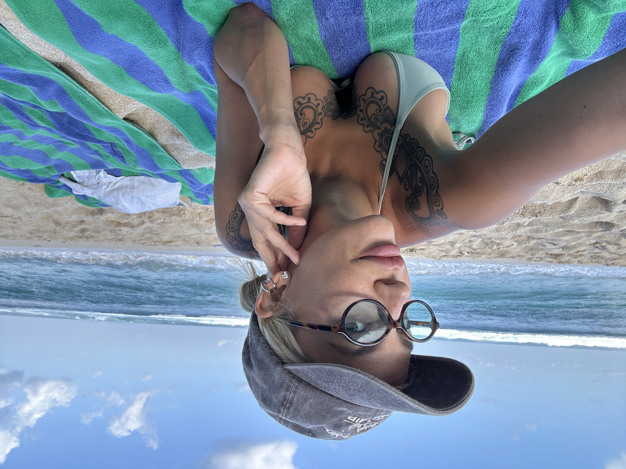 An overhead, slightly inverted perspective shows a woman relaxing on a striped beach towel (blue and green) on a sandy beach in Kuta Selatan, Indonesia. She is lying on her back, facing upwards towards the camera, with the ocean and sky visible behind her head.
The woman has light-colored hair, part of which is pulled back, and is wearing a dark grey baseball cap backwards. She has large, round, dark-rimmed glasses perched on her nose. A small, heart-shaped ear cuff or earring is visible on her right ear. Her lips are slightly parted. She is wearing a light-colored bikini top, and prominent, ornate dark-inked tattoos, possibly floral or paisley designs, adorn her upper chest and shoulders. Her left hand is raised, with fingers gently touching her face near her ear.
Behind the woman, the sandy beach transitions into the clear blue ocean with gentle white waves breaking near the shore. Beyond the waves, a clear blue sky with scattered white clouds is visible. The scene suggests a bright, sunny day. To her side, a crumpled piece of white fabric is on the sand. On the bill of her cap, there appears to be some white text or a logo, but it is obscured and not clearly legible.
