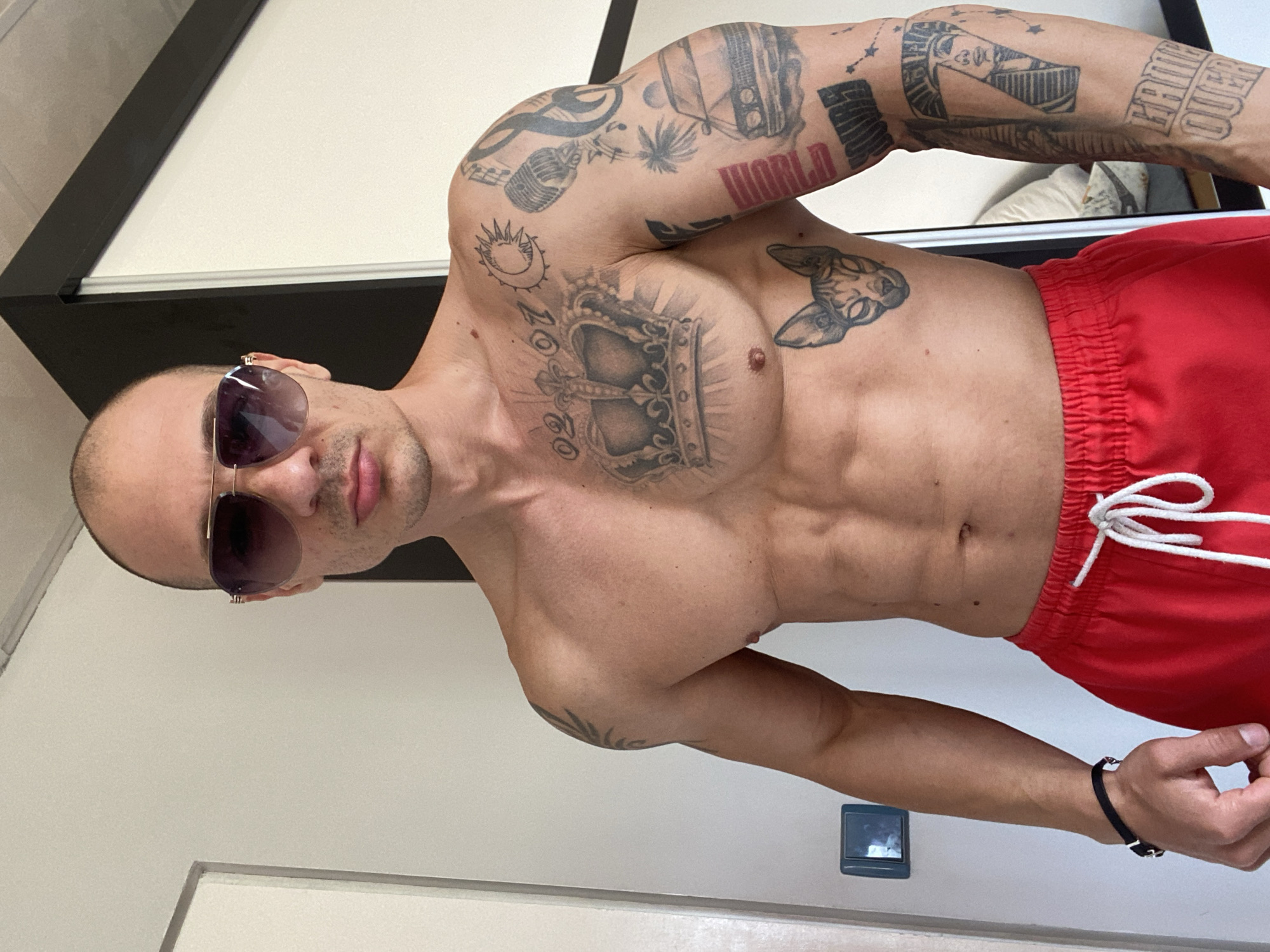 A man stands shirtless in red swim trunks. He is wearing sunglasses and a black bracelet. Tattoos are visible on his arms and chest. The location cannot be determined to be in Pleven, Bulgaria from the visual information provided.