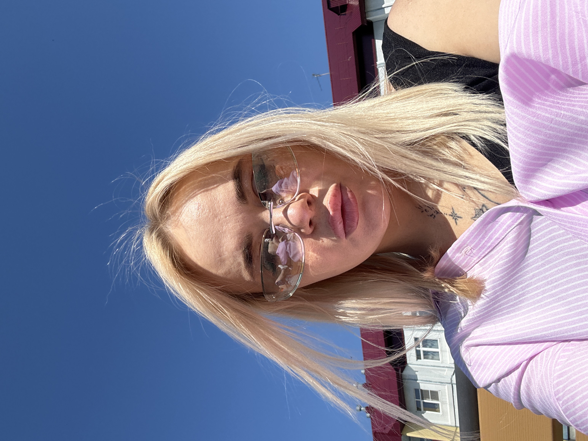 The image is a portrait of a person, likely a young woman, with long blonde hair, captured outdoors under a bright blue sky. The photo appears to be taken from a low angle, looking upwards, or the image itself is rotated 90 degrees counter-clockwise.
The subject is looking directly at the camera with a neutral or slightly pouty expression. She is wearing glasses with clear lenses and thin frames. Her hair is blonde and falls around her face and shoulders, with individual strands visible against the sky. She is dressed in a black top, overlaid with a light purple shirt featuring thin white stripes. A small tattoo, resembling a star or compass design with additional line work, is visible on her right collarbone or shoulder area.
In the background, under the clear blue sky, parts of buildings are visible. A red-colored structure or roof is partially seen in the upper left of the frame (when oriented correctly), and a lighter-colored building with multiple windows is visible towards the lower right. The bright sunlight creates strong highlights on her hair and skin, indicating a clear, sunny day.
No discernible text is visible within the image, apart from the small, stylized tattoo which is not legible.