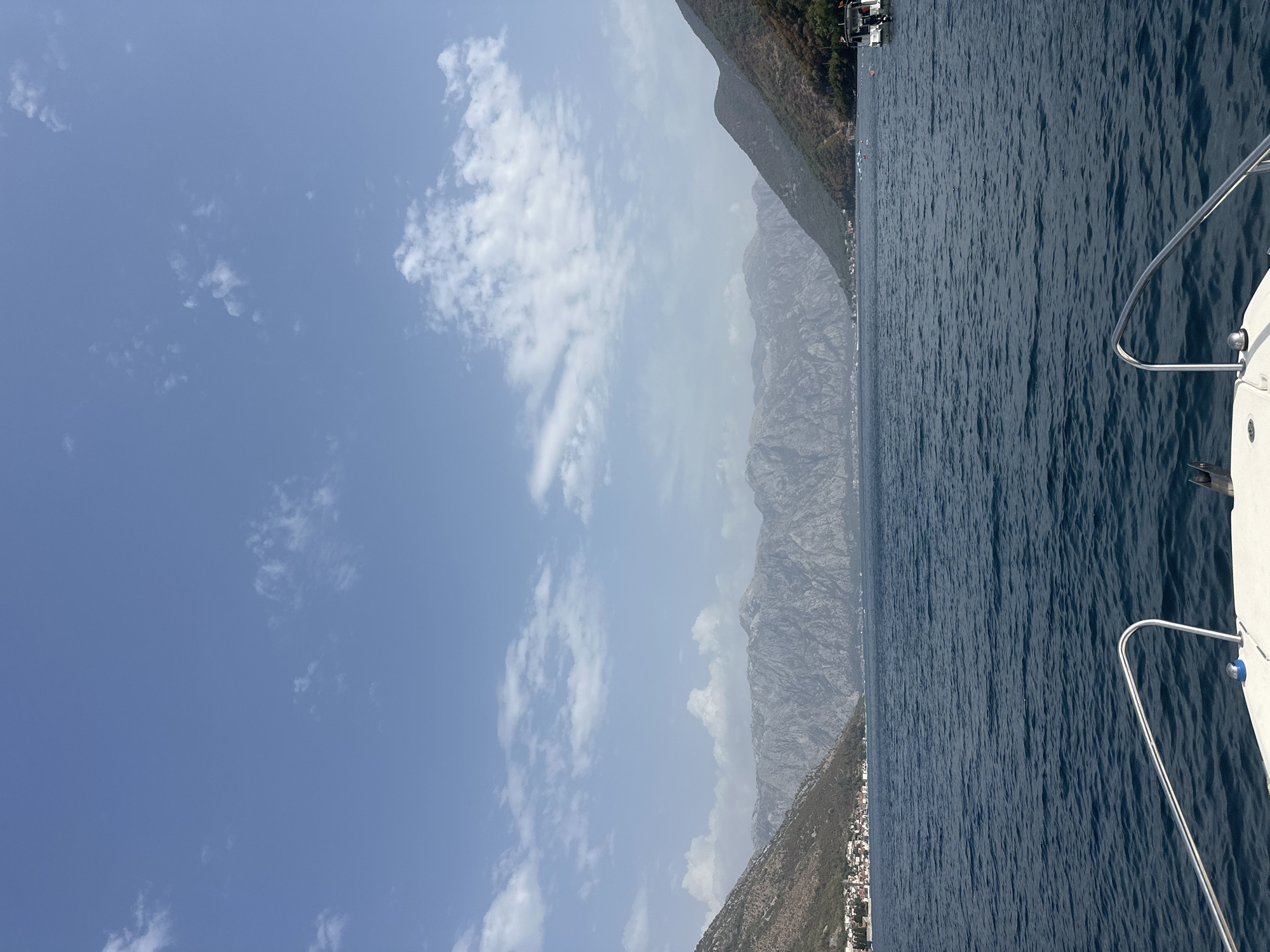 The image depicts a view from a boat's bow, looking out across a body of water towards a town nestled at the base of a mountain range. The water is a deep blue, with gentle ripples. On the left side of the frame, a town with white buildings is visible, situated on a coastline that slopes upwards into a rocky, green mountain. In the distance, a large, rugged mountain range stretches across the horizon, its peaks hazy with distance. The sky is a clear blue with scattered, wispy white clouds. The right side of the frame shows another mountainous shore and a small boat with a white hull and dark trim anchored in the water. The scene appears to be set in the Bay of Kotor, likely near the town of Perast, Montenegro, given the characteristic landscape of steep mountains rising directly from the water and the presence of a historic coastal settlement.
