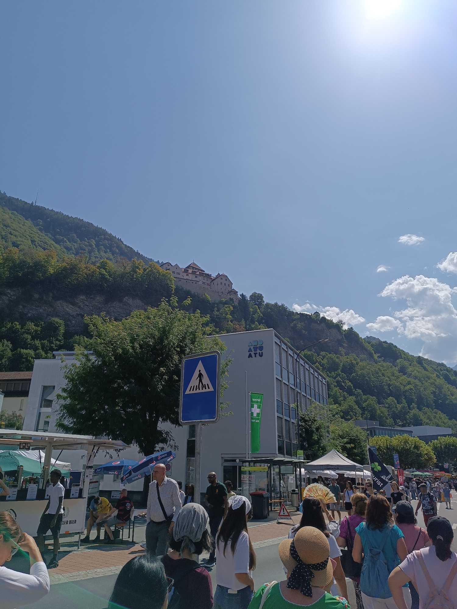 The image captures a sunny day in Vaduz, Liechtenstein, with the prominent Vaduz Castle visible atop a forested hill in the background. In the foreground, a street is filled with people walking and browsing. There are several market stalls and food vendors set up along the street. A modern, multi-story building with the logo "AGU ATU" and a "Sanität" (First Aid) banner is on the right side of the street. A pedestrian crossing sign is also visible. The sky is clear blue with some fluffy white clouds, and the sun is shining brightly, creating lens flare in the upper right portion of the image. The overall scene depicts a lively public gathering or event in the city.