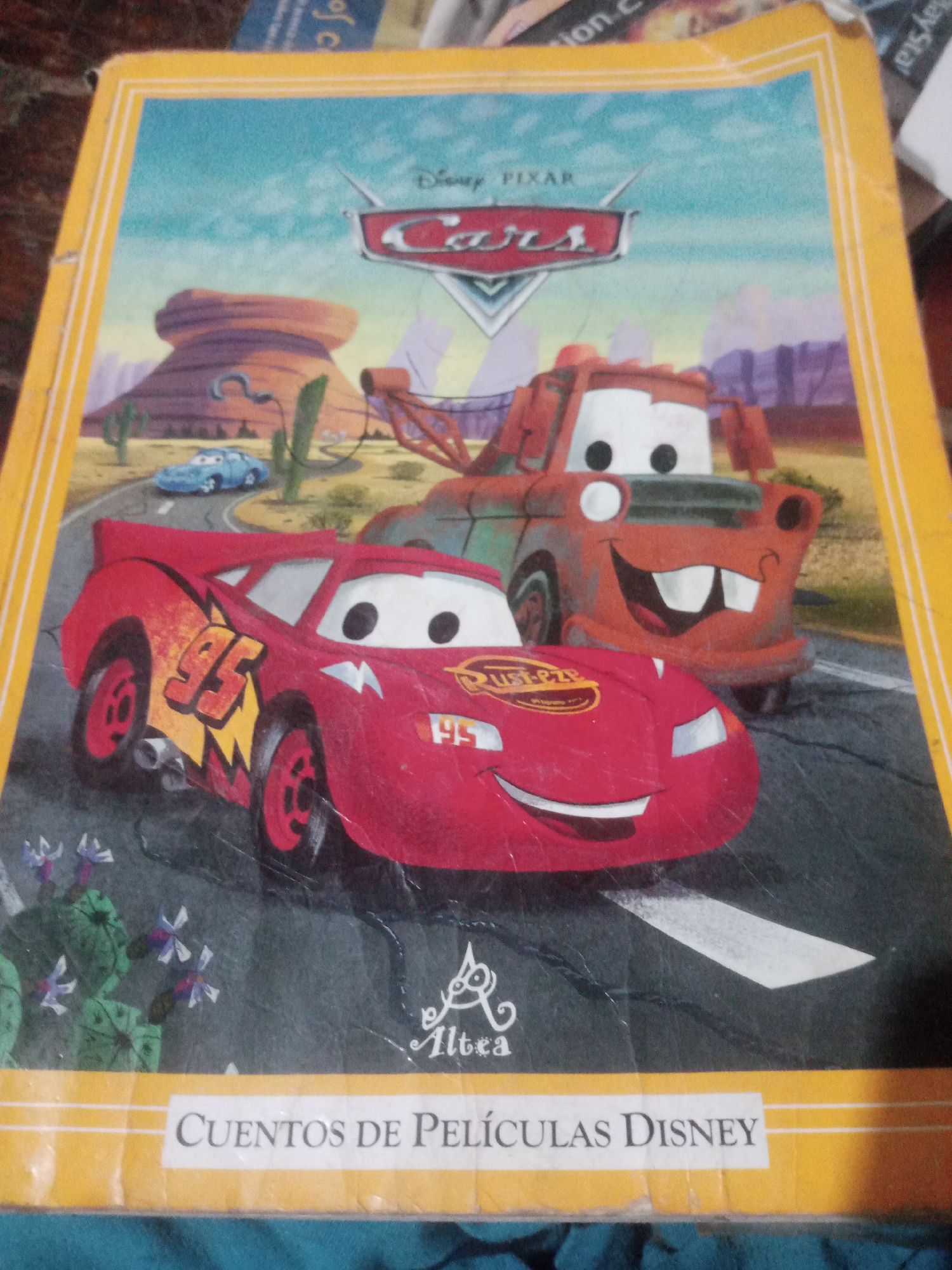 The image is a close-up, eye-level shot of the cover of a children's book titled "CUENTOS DE PELÍCULAS DISNEY" (Stories from Disney Movies). The cover features characters from the Disney Pixar movie "Cars." In the foreground, on the right, is a red race car, identified as Lightning McQueen, with his signature "95" decal visible on the side. To the left and slightly behind Lightning McQueen is Mater, a tow truck, also depicted with his characteristic grin. The background shows a desert landscape with rock formations, cacti, and a road. The book cover is slightly creased and worn. The image appears to be taken indoors, possibly in a home or library, and is not related to any specific location in Santa Ana, Venezuela.