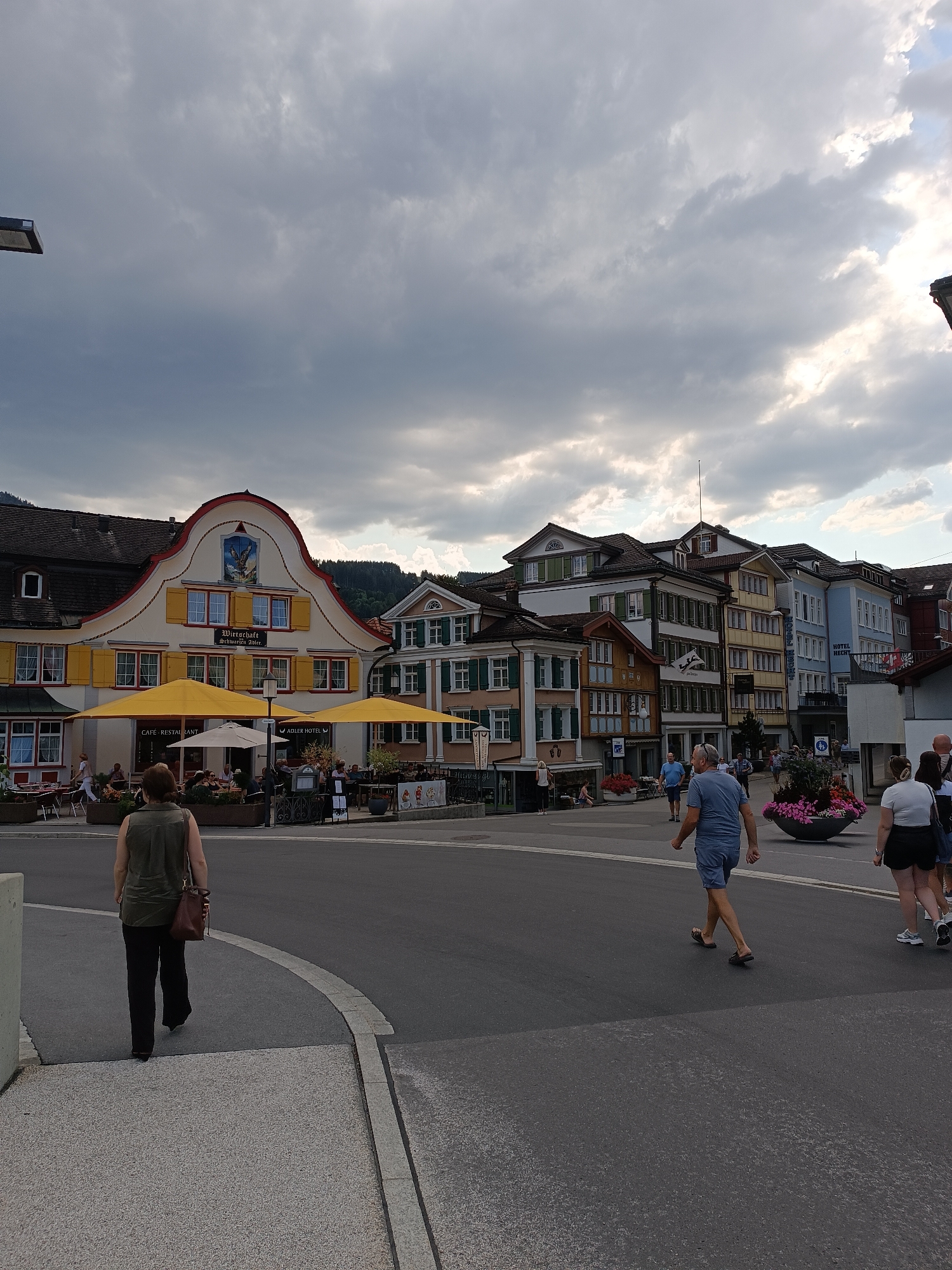 The image depicts a street scene in Appenzell, Switzerland. In the foreground, a woman with dark hair and a green sleeveless top walks away from the camera on a paved walkway. Behind her, a street opens up with several people walking and sitting at outdoor cafes. To the left, a building with a distinctive yellow and white facade, featuring a steep roof and yellow shutters, houses a cafe and restaurant. Yellow umbrellas shade outdoor seating areas. To the right, a row of colorful buildings, typical of Swiss architecture, lines the street, some bearing signs that read "Adler Hotel" and "Hotel Hecht." People are seen dining at tables and walking along the sidewalks. The sky above is cloudy, with patches of sunlight breaking through. The environment appears to be a lively town square or main street with shops, cafes, and hotels.
