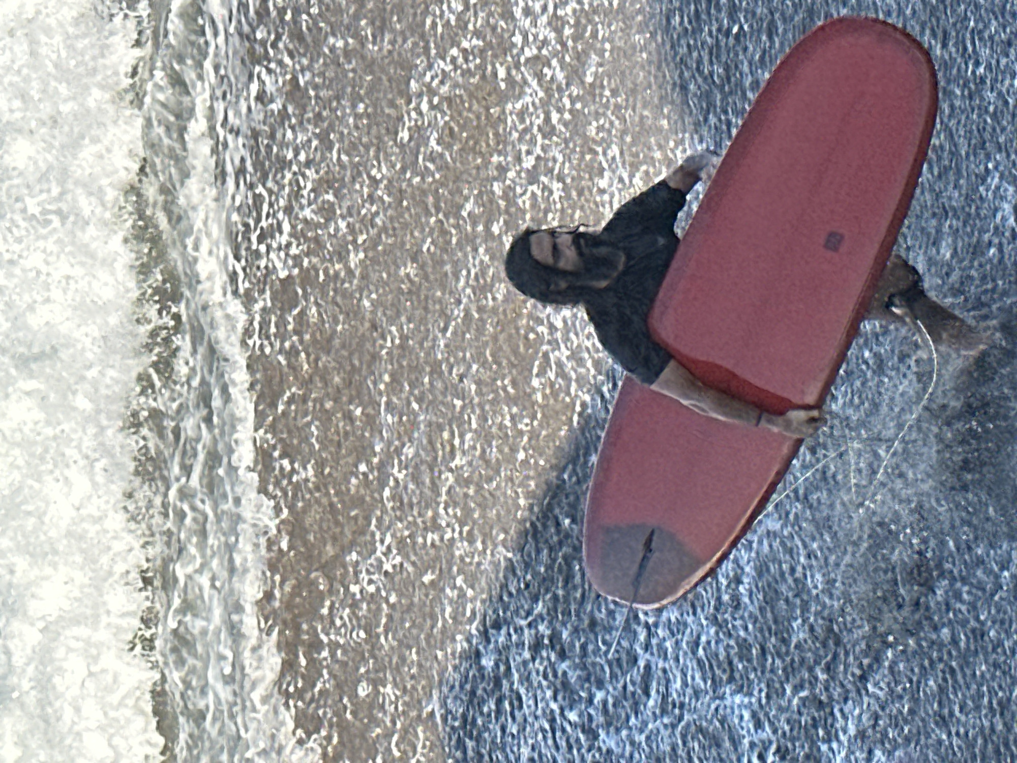 A man with dark hair and a beard, wearing a black t-shirt, walks in the water holding a red surfboard. He is in the process of entering the ocean, with waves breaking behind him and the shore visible in the foreground. The scene appears to be a beach in North Kuta, Indonesia.