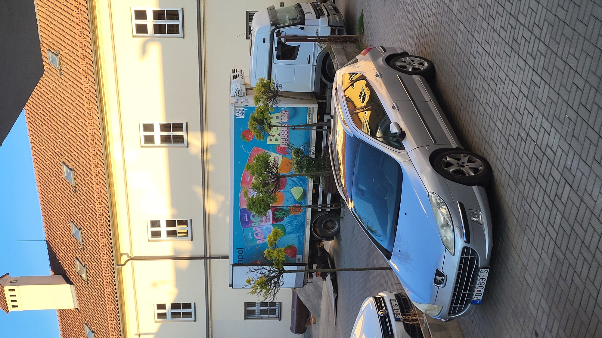 The image depicts an outdoor scene on a sunny day, likely in the city of Prešov, Slovakia, based on the license plate of the silver car. In the foreground, a silver Peugeot 407 station wagon is parked on a brick-patterned paved surface. Its license plate reads "PO 089FS". To the left of the Peugeot, partially visible, is the front of a white Volkswagen.
Behind the cars, a large white truck with a blue and yellow advertisement for "BOB BUBBLE TEA" is parked. The advertisement features colorful depictions of drinks and fruit. The truck has a refrigerated unit at the back. Several small, manicured trees with green foliage are planted in front of the truck.
In the background, a pale yellow building with a terracotta-tiled roof is visible. The building has several windows, some with white frames. A tall, light-colored chimney with horizontal vents rises from the roof. The setting appears to be an urban area, possibly a commercial or industrial zone, given the presence of the truck and building. The lighting suggests it is either early morning or late afternoon, with long shadows cast across the scene.