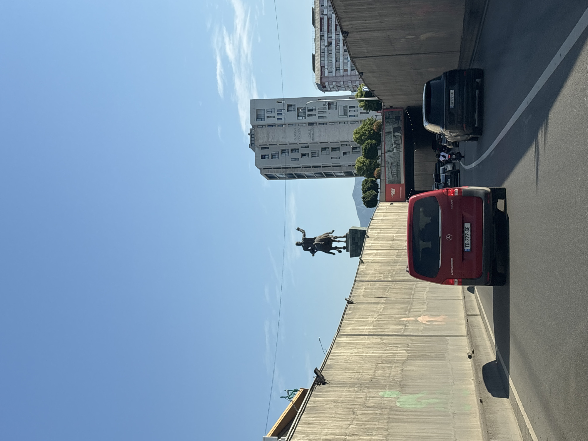 This image captures a street scene in Tbilisi, Georgia, likely near the area of Freedom Square, given the presence of the equestrian statue of St. George. The perspective is from a vehicle driving on a multi-lane road that appears to be entering or exiting a tunnel or underpass.
Dominating the upper left quadrant is the sky, a clear, bright blue with a few wispy white clouds. Below the sky, on a raised concrete structure, stands a bronze equestrian statue of a man in regal attire, his right arm raised in a gesture of command. The statue is silhouetted against the sky. To the left of the statue, a portion of a modern, multi-story building is visible.
The foreground and mid-ground are occupied by vehicles. A red Mercedes-Benz Vito van is in the left lane, its rear clearly visible with license plate "TB-777-SE". To its right and further down the road, a black Range Rover is in the right lane, its license plate reading "JU-572-UK". Other vehicles and a motorcycle are visible in the distance, suggesting moderate traffic.
The sides of the road are lined with tall, textured concrete walls, characteristic of urban infrastructure like tunnels or elevated roadways. Faint, green and pink artistic renderings, possibly graffiti, are visible on the left wall. Trees and buildings are seen in the background, with a prominent, tall, modern apartment building with numerous windows and balconies situated to the right of the equestrian statue. Another building, with a reddish facade and balconies, is partially visible behind it.
The lighting suggests it is daytime, with the sun casting shadows on the road, indicating a high sun position. The overall atmosphere is urban and active, with the infrastructure and vehicles pointing to a busy city environment.