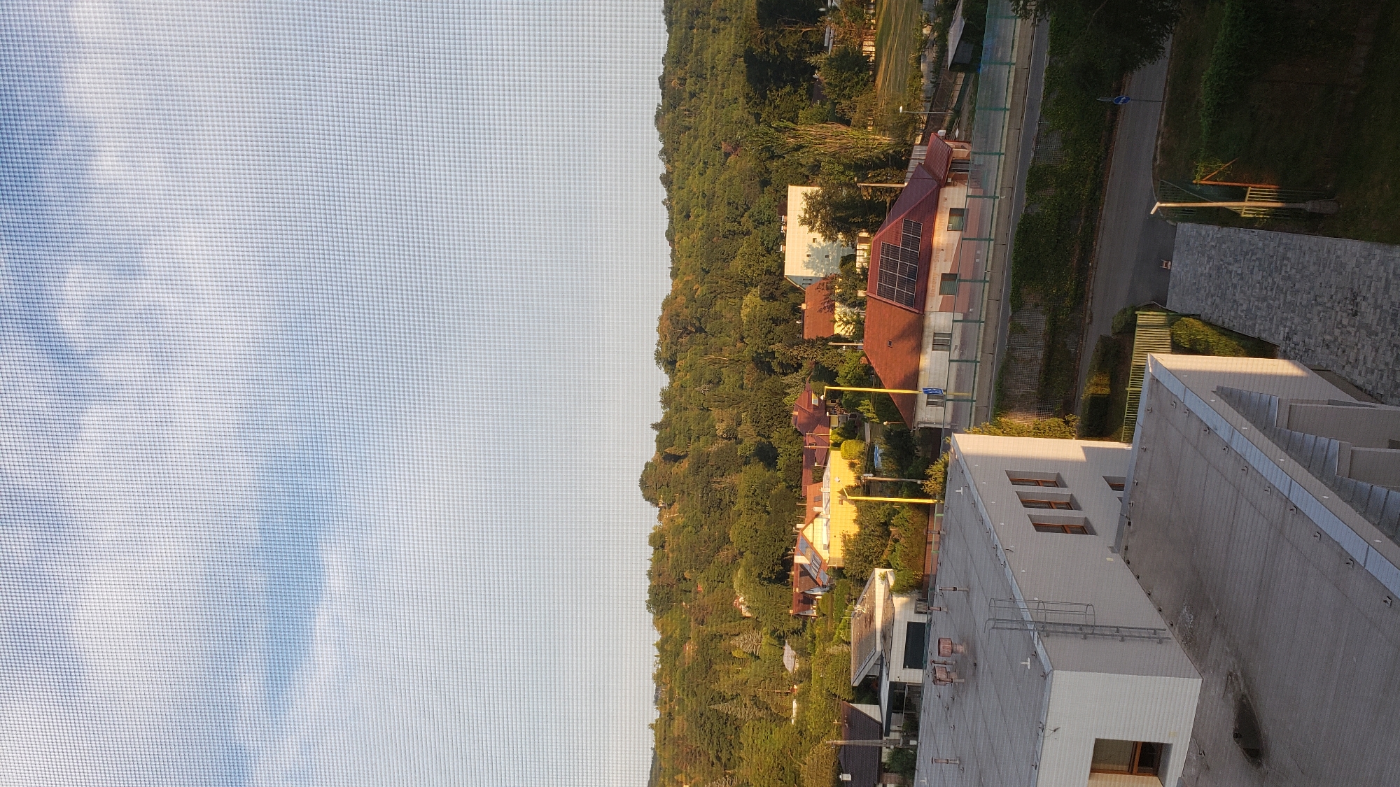 The image depicts a view of a residential area with houses and trees, seen through a window screen. In the foreground, there is a modern white building with windows and a rooftop. In the middle ground, several houses with red-tiled roofs are visible, some equipped with solar panels. A road runs through the scene, with a green fence and vegetation lining its side. Beyond the houses, a dense forest covers a hill. The sky above is partly cloudy, with a clear blue sky visible in some areas. The scene appears to be a suburban or semi-rural setting, likely taken during daylight hours. The location is Prešov, Slovakia.