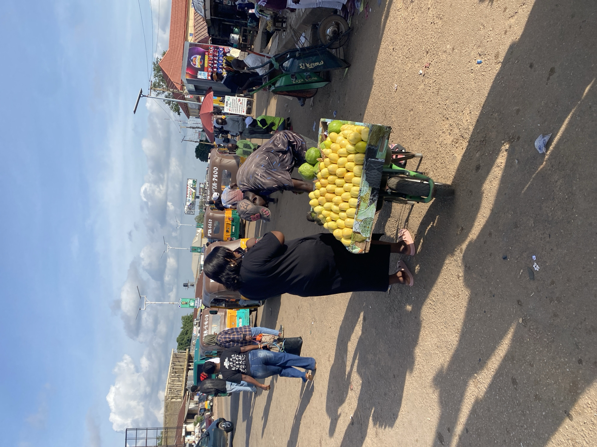 In Gura Top, Nigeria, a street scene unfolds under a bright, cloudy sky. In the foreground, a woman in a black dress and pink sandals stands by a cart laden with bright yellow produce, likely fruits, arranged in neat rows. To her left, a man wearing a patterned shirt, dark pants, and a baseball cap leans over the cart, appearing to arrange or inspect the goods. Behind them, several auto-rickshaws, known locally as Keke Napeps, are parked or moving, their colorful bodies contrasting with the dusty road.
A woman in a black t-shirt with a graphic print and blue jeans walks away from the camera, carrying a black bag. Another person in a plaid shirt and headscarf walks alongside her, holding a patterned bag. In the background, various buildings line the street, including one with a red tiled roof and another under construction. Banners and signs are visible, one advertising a "Festival." The ground is a mixture of dirt and gravel, with long shadows cast by the people and vehicles, indicating a sunny day. The overall impression is of a bustling marketplace or a busy street corner in a Nigerian city.