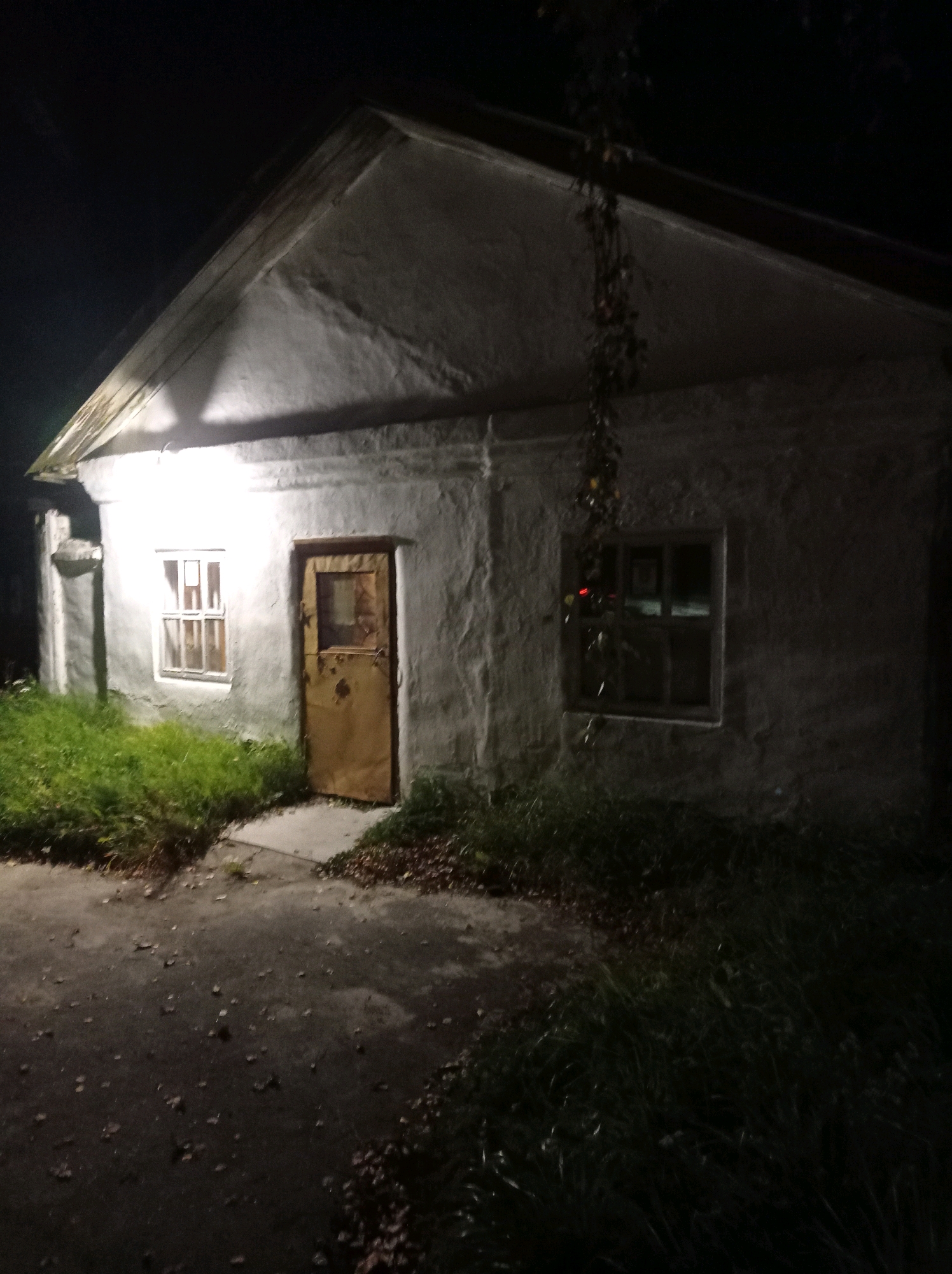The image shows a low-angle, night-time exterior view of a small, dilapidated building. The building has a stuccoed exterior, a gabled roof with exposed wooden beams, and at least two windows. One window, to the left of the entrance, is illuminated from within and has a grid pattern. The entrance features a single, heavy, weathered metal door with a small square windowpane. Overgrown green grass and fallen leaves are visible around the building. A vine hangs from the roofline above the door. The scene appears to be situated in an undeveloped or neglected area, potentially on the outskirts of Degtyarsk, Russia, given the overall condition of the structure and its surroundings. The lighting suggests artificial illumination, possibly from a streetlamp or the building's interior, casting shadows and highlighting the rough texture of the walls.