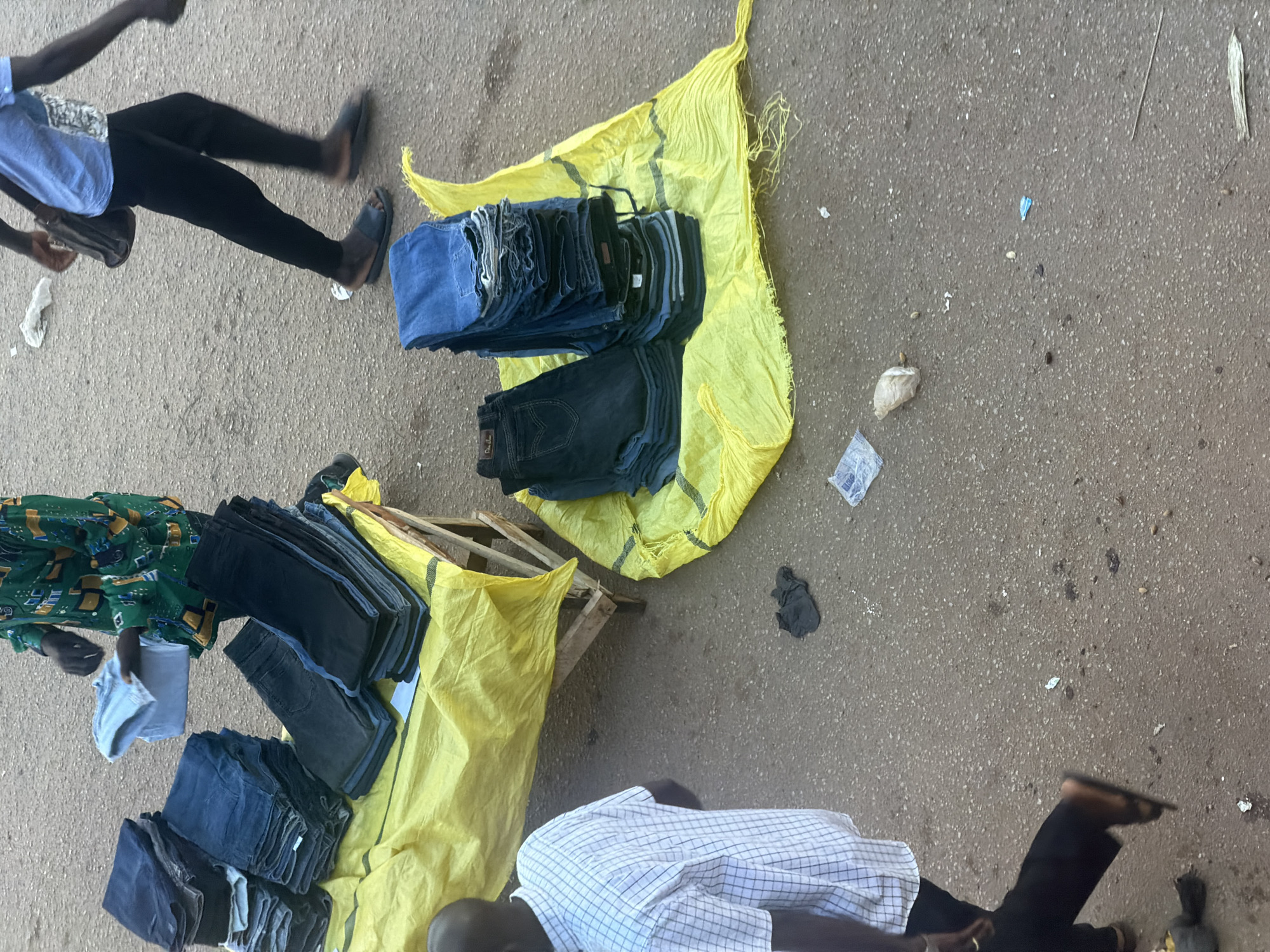 This media file captures a scene on a dusty, paved ground, likely a street or market, in Bukuru, Nigeria. The image features at least three individuals engaged in what appears to be a commercial transaction or preparation for one.
One subject, wearing a light blue shirt and dark trousers, is walking away from the camera, holding a small, dark bag. Their motion suggests they are in transit. Another individual, dressed in a green patterned garment, is seen from above, interacting with a stack of light blue folded clothing, possibly T-shirts. Their hands are engaged in handling the garments. A third person, wearing a checkered shirt and dark trousers, is lying down, partially visible at the bottom right of the frame, with their legs stretched out.
Dominating the scene are stacks of folded denim jeans, primarily in dark blue and black hues. These are neatly piled on a yellow tarp and a wooden pallet, suggesting an outdoor stall or display of merchandise for sale. A portion of a green and yellow patterned fabric, possibly part of another item of clothing or textile, is also visible. The overall setting is informal and appears to be outdoors, with the ground showing signs of wear and scattered debris, including small white and blue plastic fragments, and a piece of white paper.
The lighting suggests it is daytime, likely mid-morning or afternoon, as there are no strong shadows, and the scene is generally well-lit. The weather appears to be dry, given the dusty ground. The atmosphere seems bustling and typical of a local market environment. No visible text is present in the image.