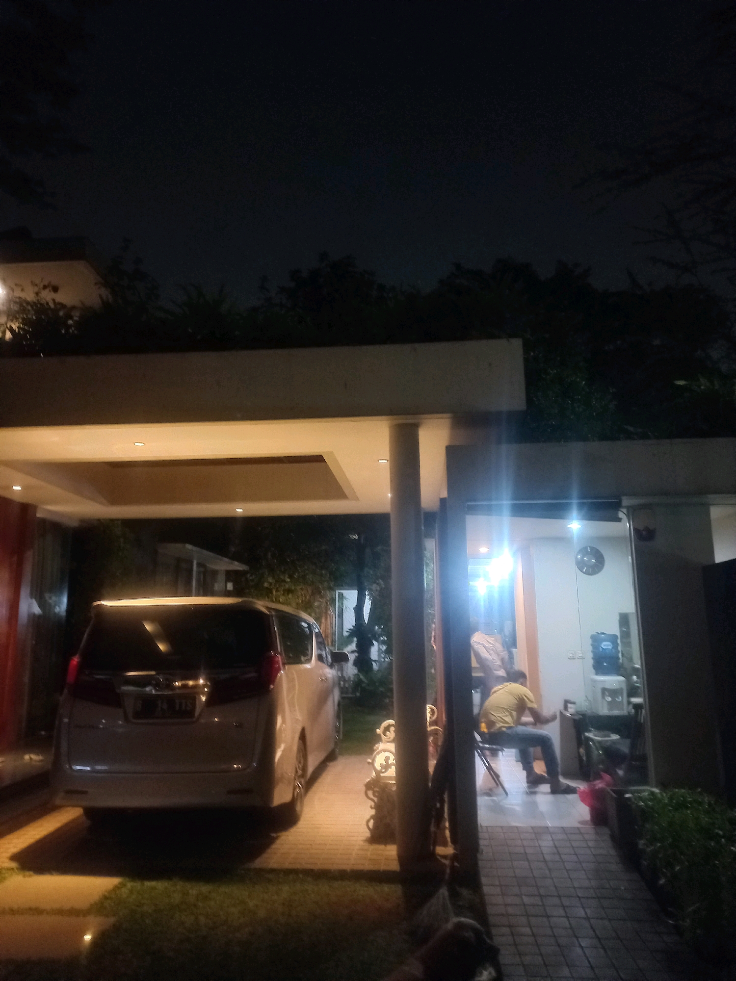 The image depicts a nighttime scene in South Jakarta, Indonesia, likely a residential area or a commercial establishment. In the foreground, a light-colored minivan is parked under a covered carport. The minivan has its taillights on and a license plate is visible. To the right of the minivan, an open doorway reveals an interior space with a person wearing a yellow shirt and blue pants sitting on a chair. Inside, there's a water dispenser, a clock on the wall, and what appears to be a kitchen or storage area. The ground is paved with interlocking bricks. Trees and foliage are visible in the background, adding to the outdoor ambiance. The scene is illuminated by artificial lighting, creating a somewhat grainy and dark atmosphere characteristic of nighttime photography.