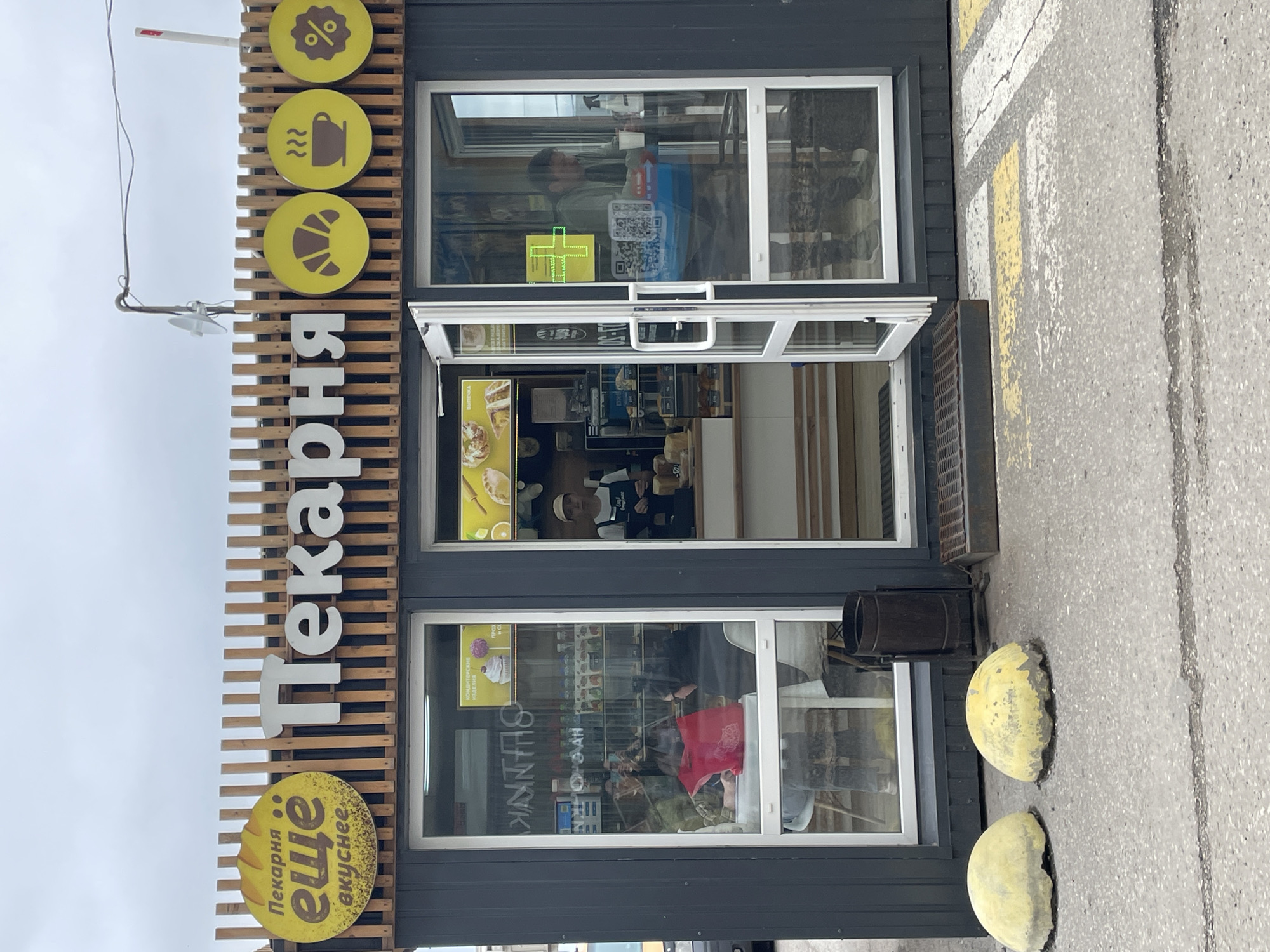 The image shows a small bakery storefront in Kirov, Russia. The bakery is named "Пекарня ещё вкуснее" (Bakery even tastier). The storefront features a large sign with the bakery's name and a series of yellow circular icons depicting a croissant, a cup of coffee with steam, and a percentage sign. The main entrance has a glass door that is propped open. Inside the bakery, a person wearing a white cap and apron is visible behind a counter. Shelves with baked goods are visible through the windows. In front of the bakery, there are two concrete bollards painted yellow. The surrounding area is paved with asphalt and has yellow and white road markings. The sky is overcast.