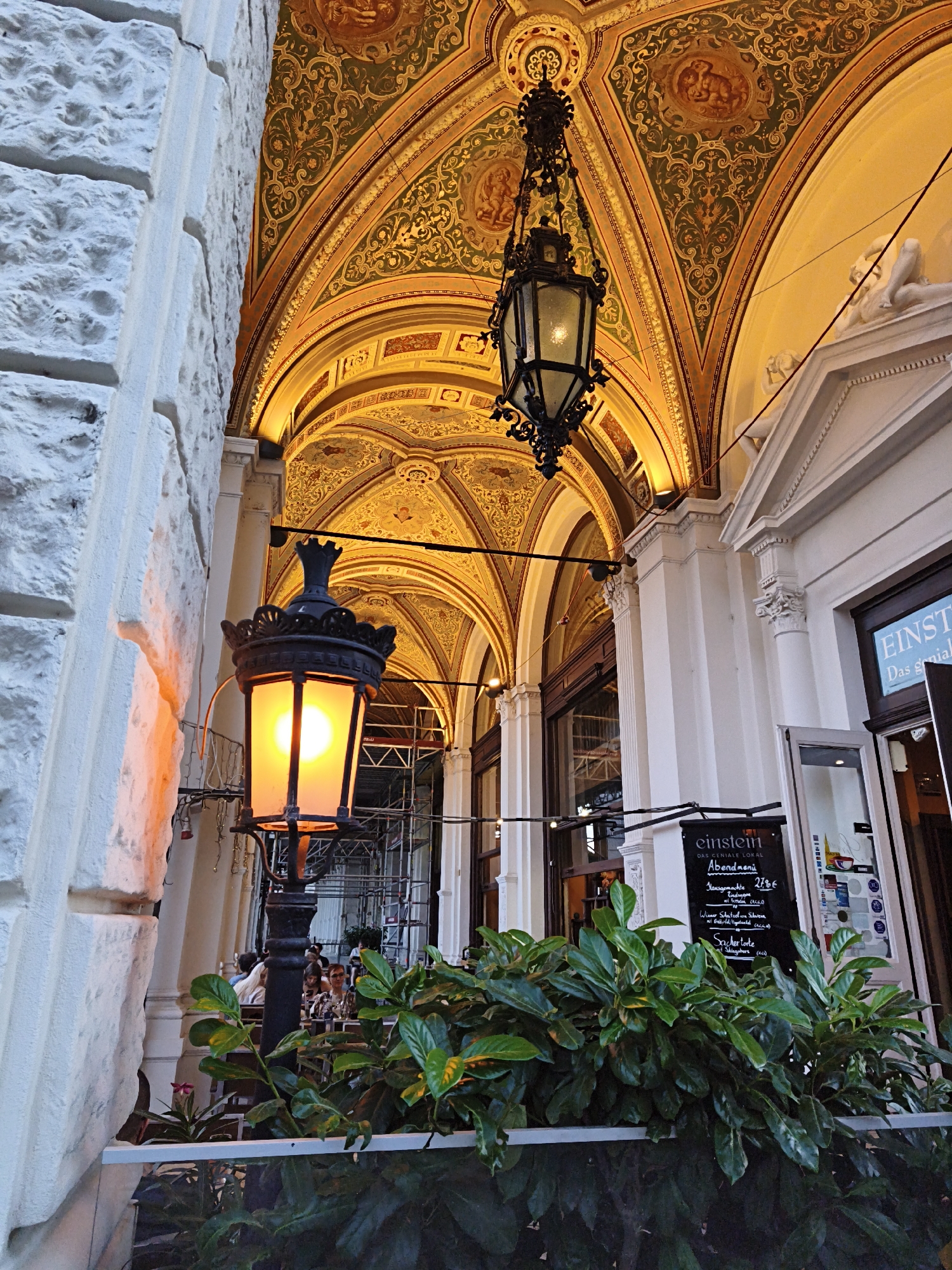 The image depicts an outdoor seating area of a Viennese establishment, likely a cafe or restaurant, characterized by its ornate architecture. The ceiling of the covered walkway features elaborate gold and green decorative frescoes and arches. A black, ornate hanging lantern is suspended from the ceiling, while a taller black lamp post with a glowing orange light stands closer to the viewer.
In the background, patrons are seated at tables, partially obscured by lush green foliage in the foreground. Scaffolding is visible behind the seating area, suggesting ongoing construction or renovation. A sign with the name "Einstein" and menu details is mounted on the wall near the entrance to the establishment. The building facade exhibits classical architectural elements with columns and sculpted details. The overall impression is of a sophisticated European urban setting.