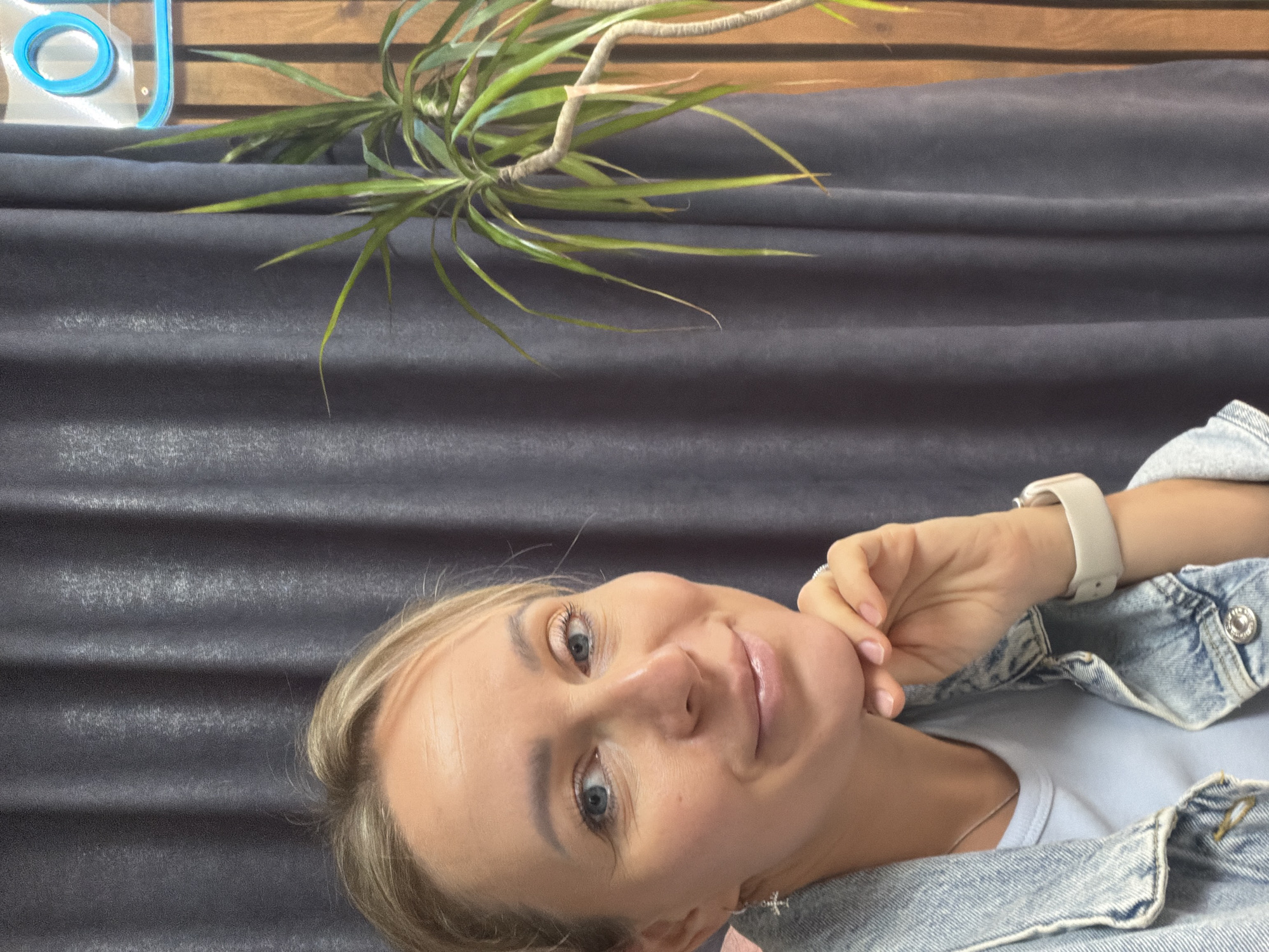 A casual, close-up portrait shows a woman with blonde hair and light blue eyes, looking directly at the camera with a soft smile. Her head is tilted, and her right hand is gently touching her chin. She is wearing a light blue t-shirt layered under a light-wash denim jacket. A white smart watch is visible on her left wrist, and a small, cross-shaped earring adorns her left ear.
The background features a dark, textured fabric, likely a curtain or upholstered surface, running horizontally behind her. Above this, a wooden wall or panel is visible. A green, leafy plant, possibly a Dracaena, extends from the left across the upper part of the background. In the top left corner, a partial view of a blue, neon-like light fixture, appearing as a circle or the letter "O," is present. The lighting is soft and even, suggesting an indoor setting. The image captures a serene and friendly expression from the woman. No specific activity or event other than posing is depicted. No discernible text is visible in the frame. The image is rotated, with the subject's head oriented towards the right side of the frame.