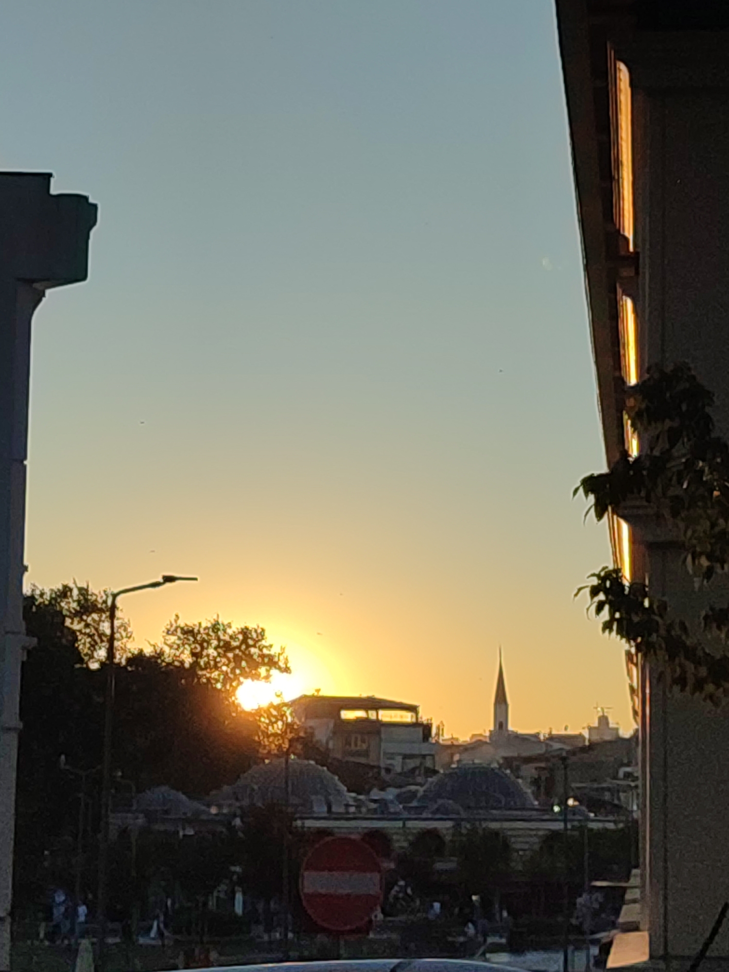 This image depicts a sunset over the Üsküdar district of Istanbul, Turkey, as viewed from a building. The sky transitions from a pale blue at the top to a warm orange glow near the horizon, with the sun partially obscured by trees. Silhouetted against the sunset are various buildings, including what appears to be a mosque with a dome and a tall, slender minaret. In the foreground, a large, dark structure, possibly a building or a wall, frames the right side of the image. A streetlamp is visible to the left of the frame. A red and white circular "no entry" sign is also present in the lower middle section of the image. The overall scene is tranquil, capturing the end of the day in a cityscape.