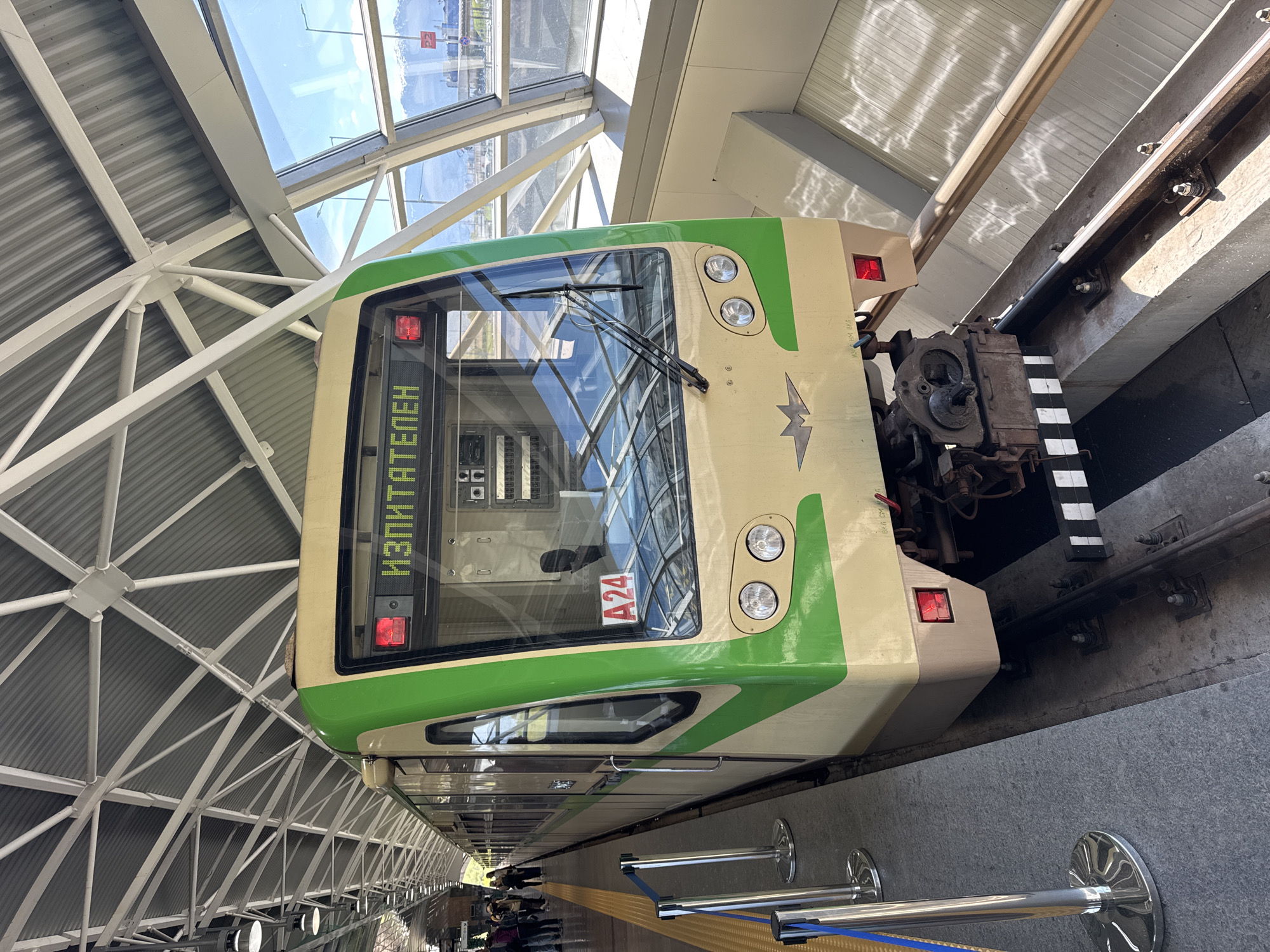 The image depicts the head of a metro train, likely located in Sofia, Bulgaria, as indicated by the Cyrillic inscription "ИЗПИТАТЕЛЕН" (Izpitatelen), which is a station in the Sofia Metro. The train is a light-colored vehicle with green accents and features a large front windshield with wipers. An "A24" marker is visible.
The train is situated within a modern metro station, with a ceiling constructed of white metal beams and panels. Outside the glass windows of the station, there is a view of the urban landscape. The platform is visible beneath the train, where people are standing, observing the scene. A line of polished chrome safety bollards connected by blue straps runs parallel to the platform edge. The train is positioned on rails, with a contrasting black and white striped structure visible on the adjacent track, possibly part of the rail mechanism.