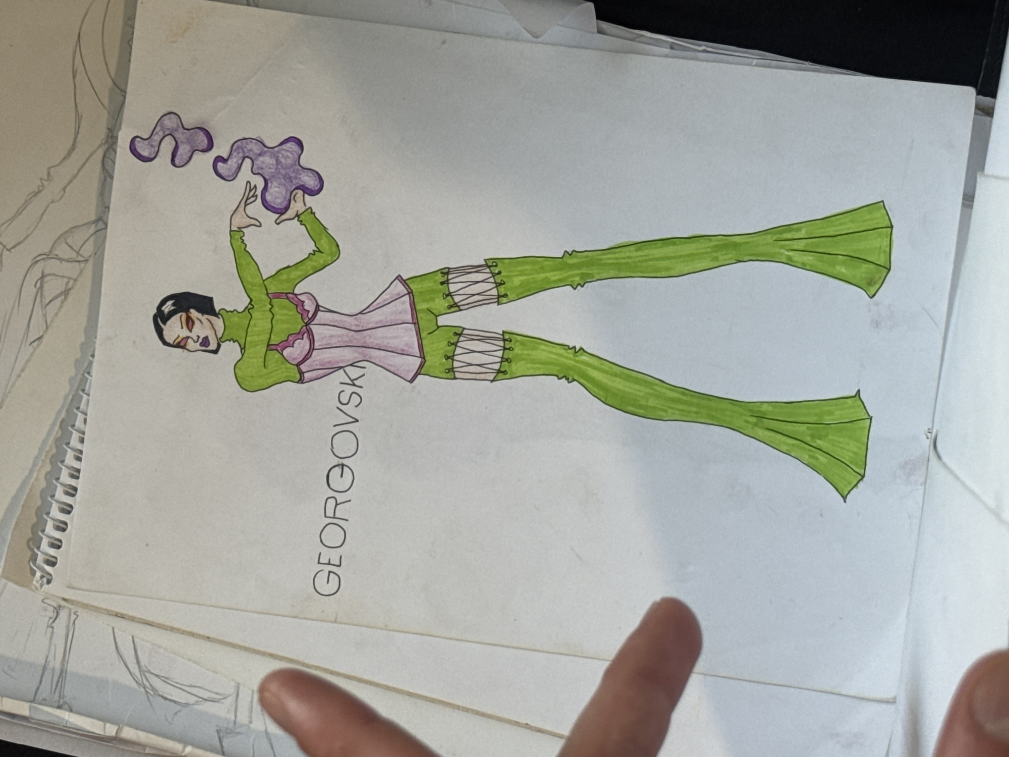 This media file shows a fashion illustration of a person in a stylized outfit. The figure is depicted vertically, with green, flared trousers and a pink corset with green accents. The person has short black hair and dark eye makeup. There are two abstract purple shapes floating above the figure's hands, which are raised as if manipulating them. The illustration is on a page that appears to be part of a sketchbook or portfolio, as suggested by the spiral binding on the left side and other sketches underneath. The name "GEORGOVSK" is written vertically along the left side of the illustration. The image is taken from a top-down perspective, with a hand visible in the lower right corner, suggesting someone is holding or viewing the artwork. The lighting is even, indicating an indoor setting, likely a studio or workspace. The overall mood is artistic and creative, showcasing a unique fashion design.
