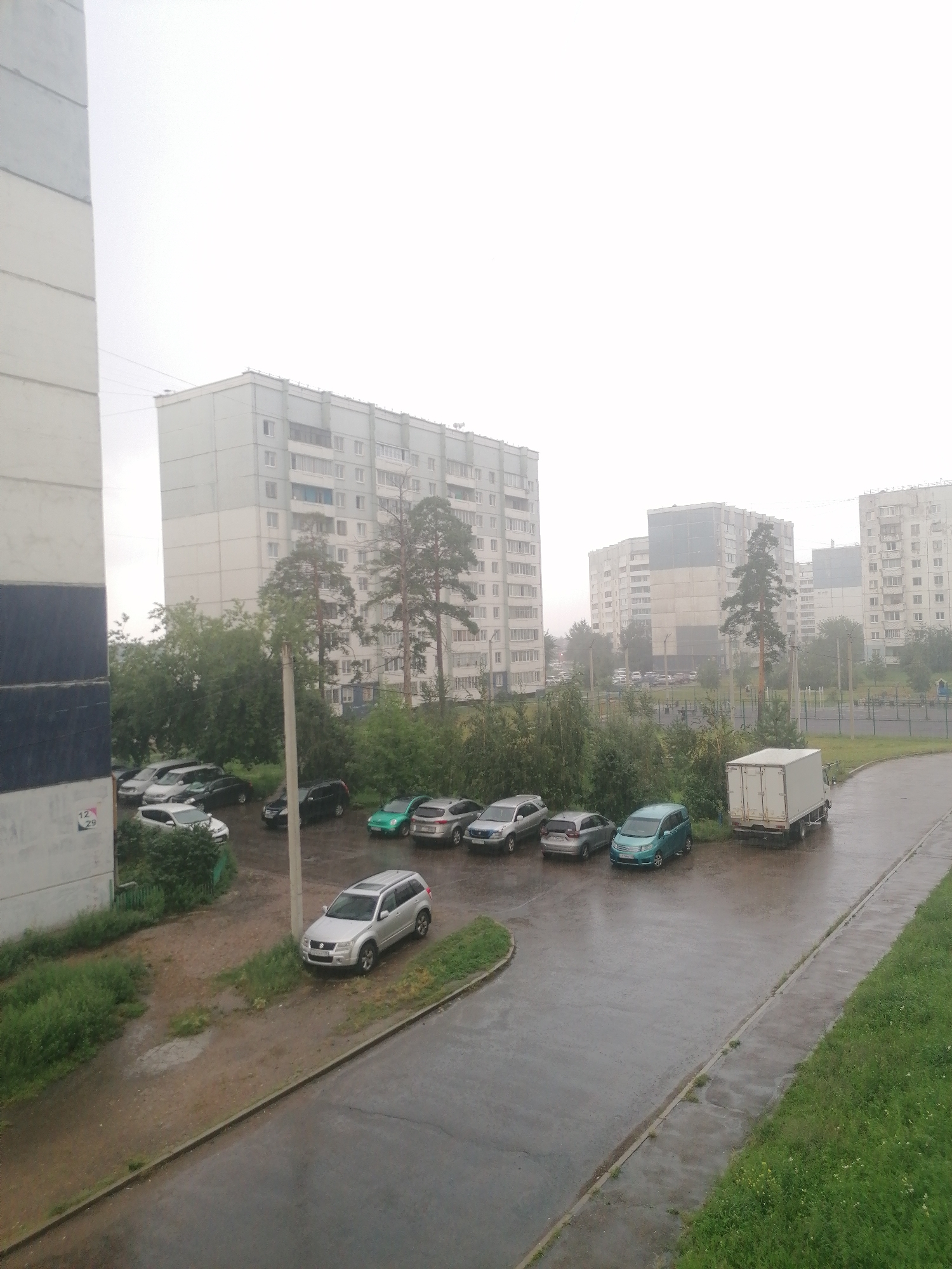The image depicts an overcast day in a residential area of Angarsk, Russia. In the foreground, to the left, is the side of a multi-story apartment building with a distinct blue and grey striped pattern. To the right, a paved road curves around a grassy area and a parking lot. Several cars are parked in the lot, including silver SUVs, a dark sedan, and a green hatchback. A white box truck is parked at the edge of the road. In the background, multiple multi-story apartment buildings are visible, characterized by their light-colored facades and numerous windows. Trees, primarily evergreens, are scattered throughout the scene, with a few taller pine trees standing out. The ground is wet, indicating recent or ongoing rain. There are no people visible in the image.