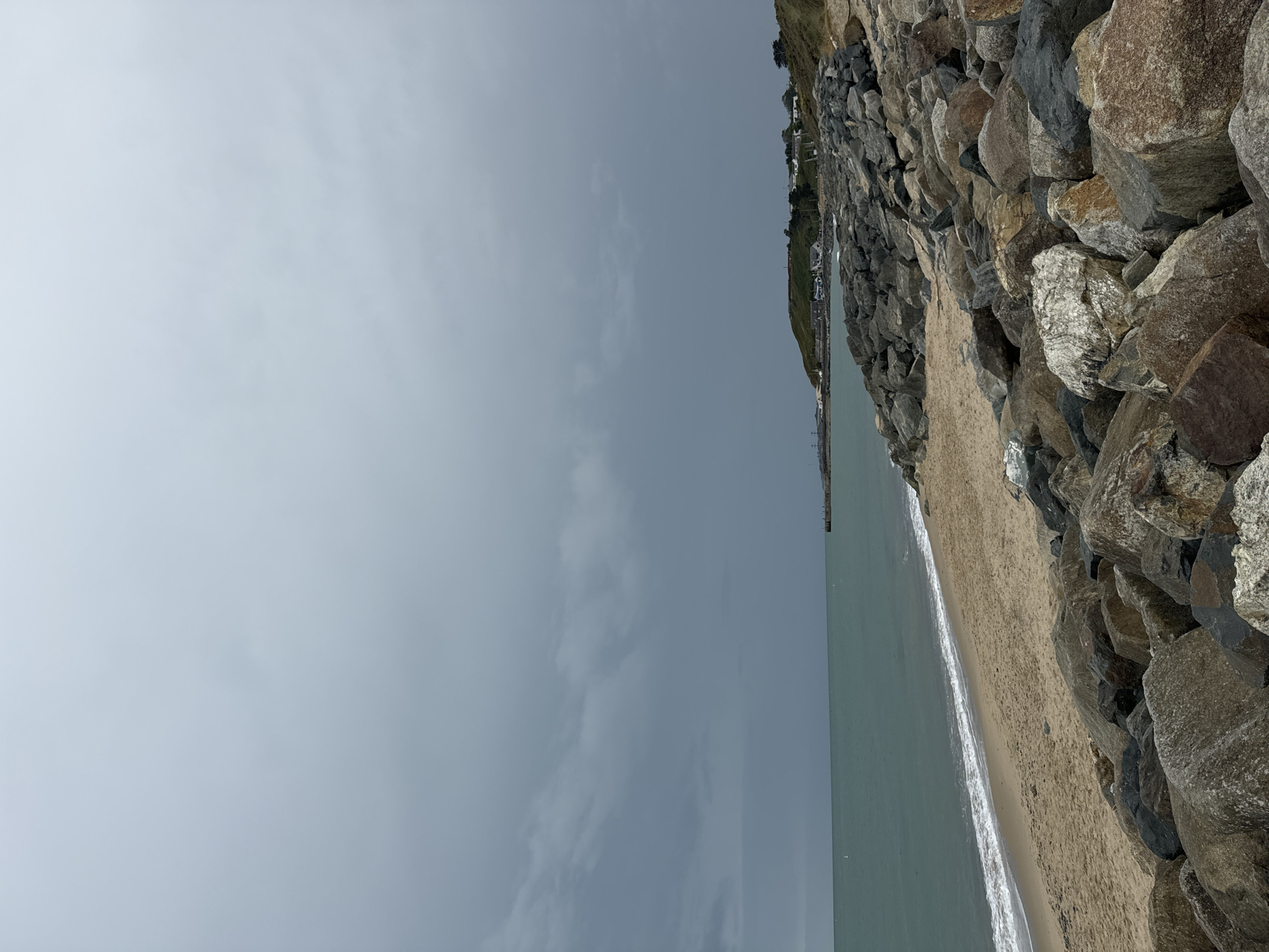 The image depicts a stony beach in Courtown, Ireland under a cloudy sky. The sea is a muted green, with gentle waves lapping onto the sandy shore. A prominent rock breakwater lines the edge of the beach, composed of large, irregular grey and brown stones. In the distance, along the coastline, a small cluster of houses can be seen nestled on a green hill. The sky is overcast with thick, dark clouds, suggesting impending rain or a storm. The overall scene is one of a rugged coastal landscape.