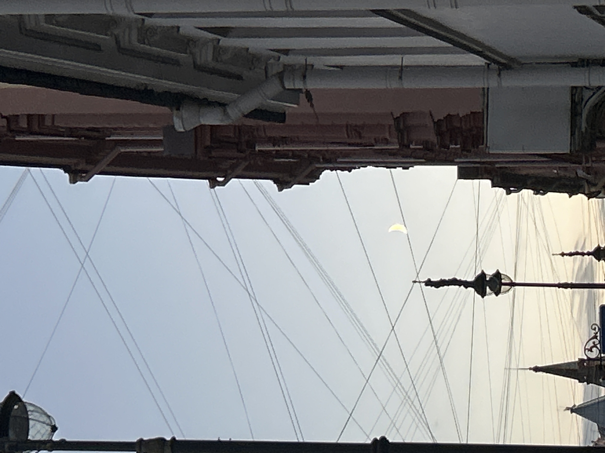 A street scene at dusk in an unknown city. The sky is a pale blue with a crescent moon visible. Multiple power lines crisscross the sky. In the foreground, on the left, is the dark pole of a street lamp. To the right, the side of a building with ornate architectural details is visible. The building is constructed of brick and has a light pink facade with white decorative elements. A white downspout runs vertically down the side of the building. In the middle ground, several street lights and spires of buildings are visible. The image appears to be taken from a low angle, looking upwards.