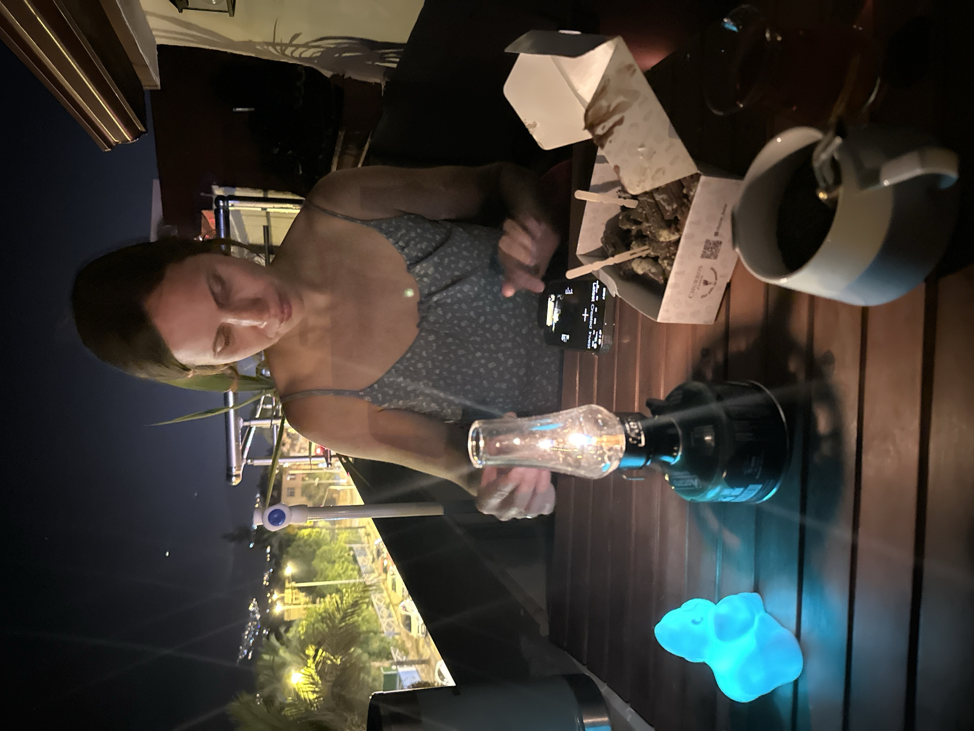 A woman is seated at a wooden table outdoors at night, with a cityscape visible in the background. She is wearing a floral print dress with thin straps. In front of her on the table is a small oil lamp that is lit, a box of churros with toothpicks, a smartphone, and a mug of coffee. To her left, a blue, glowing decorative object is visible. The scene appears to be on a balcony or terrace, overlooking Fethiye, Turkey, with distant lights and some palm trees. The overall ambiance is one of a relaxed evening meal or snack.