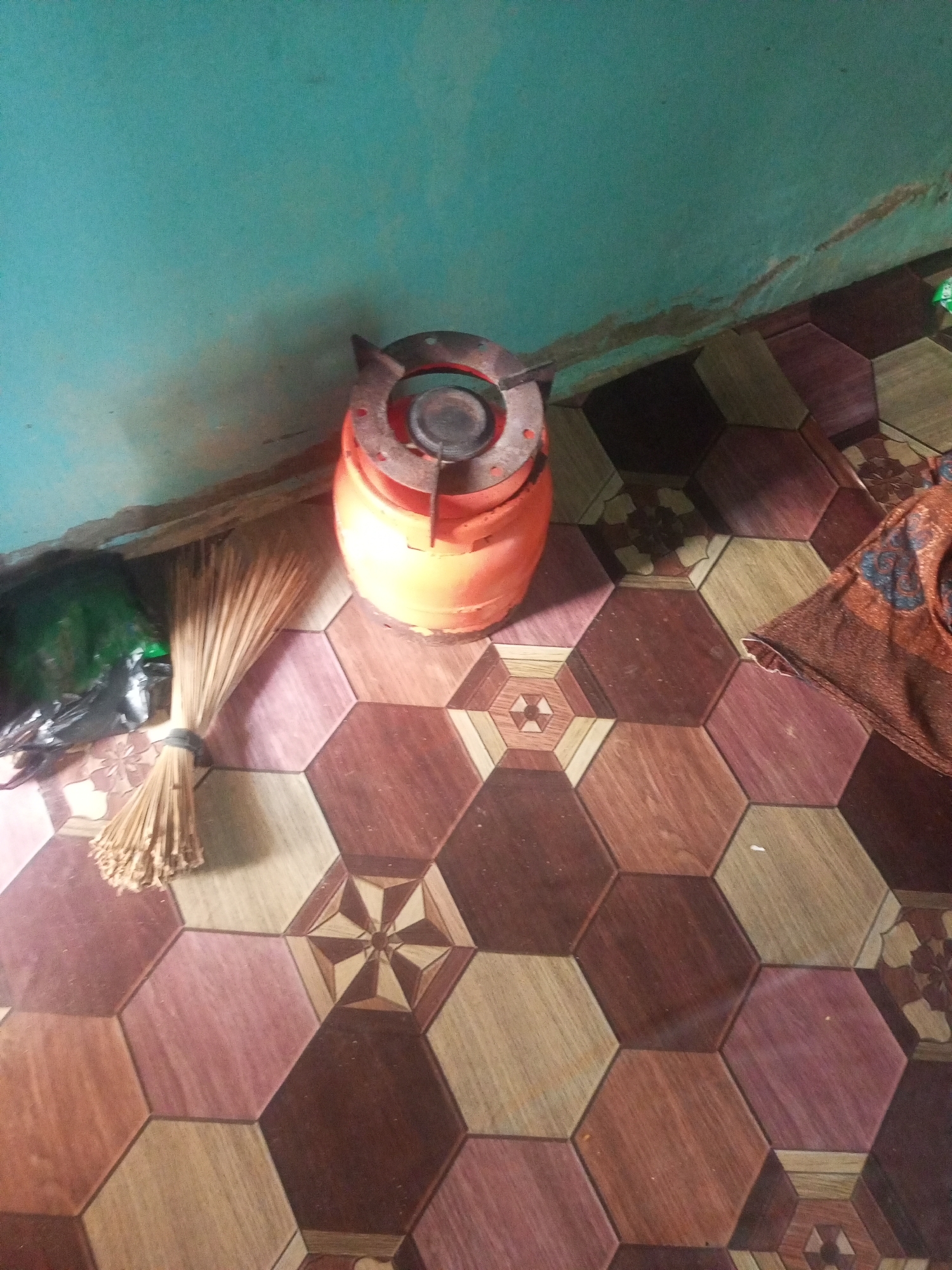 In Jalingo, Nigeria, a gas cylinder with a stovetop is positioned near a wall painted turquoise. Adjacent to the gas cylinder, a bundle of dried grass is tied with a black band. A green plastic bag lies on the floor beside the broom. The floor is covered with hexagonal tiles in various shades of brown and pink, some featuring intricate geometric patterns. A portion of a patterned fabric is visible on the right side of the frame.