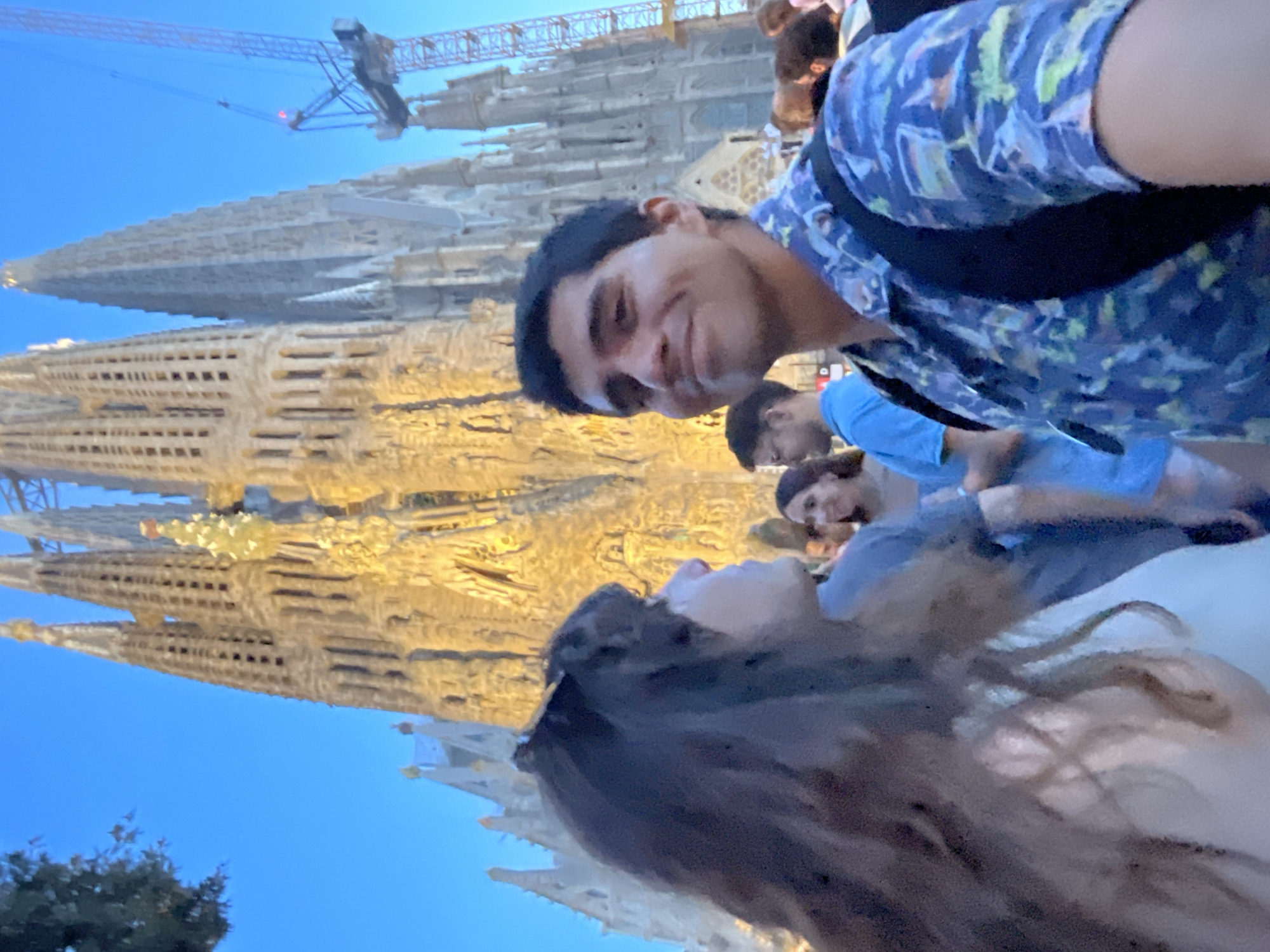 The image captures a selfie taken in front of the Sagrada Familia in Barcelona, Spain. In the foreground, a man with dark hair and a patterned blue shirt smiles at the camera. To his left, a woman with long, dark, wavy hair and sunglasses on her head is visible from the side. Behind them, other tourists are seen in the background, and a construction crane is present on the right side of the frame. The Sagrada Familia, a large and ornate basilica, is illuminated and dominates the background. The sky above is a clear, deep blue, suggesting it is late afternoon or early evening.