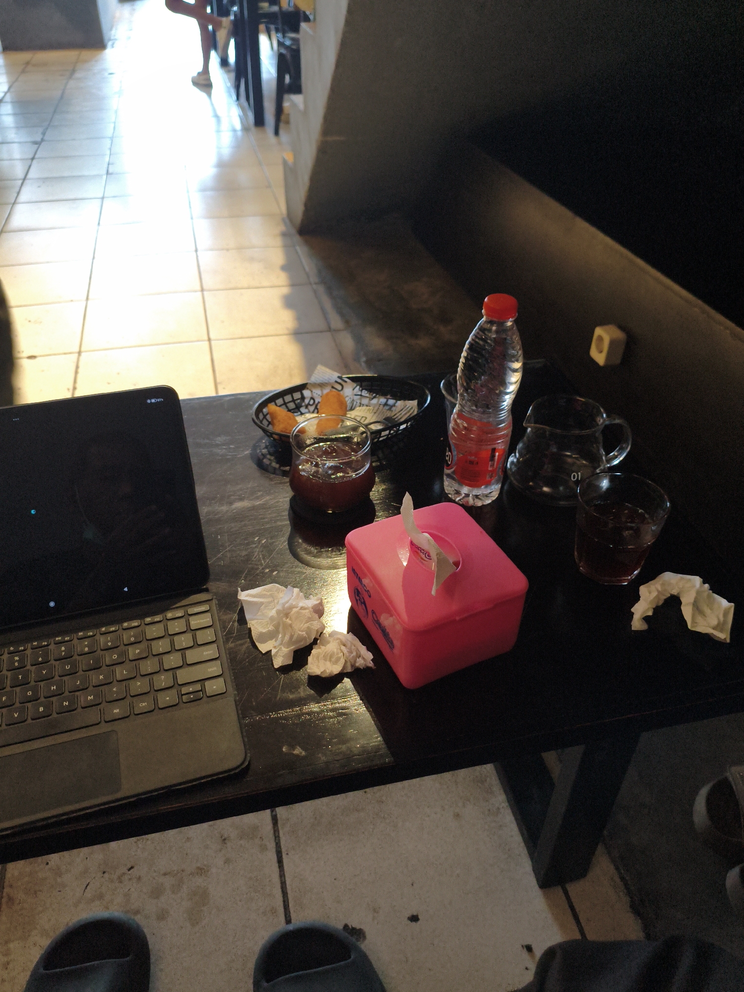 A tablet computer with a keyboard is positioned on a dark-colored table in what appears to be a cafe or restaurant. The tablet's screen is black, displaying a small blue circle. To the right of the tablet, a small black basket contains several pieces of fried food, possibly chicken nuggets. Next to the basket is a glass of dark liquid, likely a beverage. Further to the right, a clear glass pitcher sits beside a full glass of the same dark liquid. A clear plastic bottle of water with a red cap is also present on the table. In the foreground, a bright pink tissue box with a tissue emerging from it is situated to the left of the glass pitcher. Several crumpled paper tissues are scattered on the table near the tissue box and the tablet. In the background, a person's legs and feet are visible, standing on a tiled floor that reflects sunlight streaming in from the left, suggesting a well-lit indoor space. The overall scene depicts a casual setting where someone is likely working or relaxing. The location is identified as City of Tebing Tinggi, Indonesia, which implies the cafe is situated in this region.