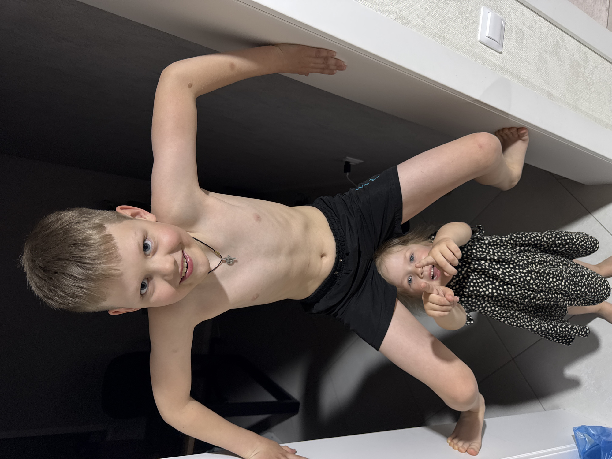 Two children are posing for a photo. A shirtless boy with blond hair is holding onto the ceiling, with a younger girl, wearing a black dress with white polka dots, standing under his legs. The background consists of a white wall, a light switch, and a dark, undefined space. It is impossible to ascertain the exact location in Gornyi (Dvurechenskoe S/P), Russia, from this image.