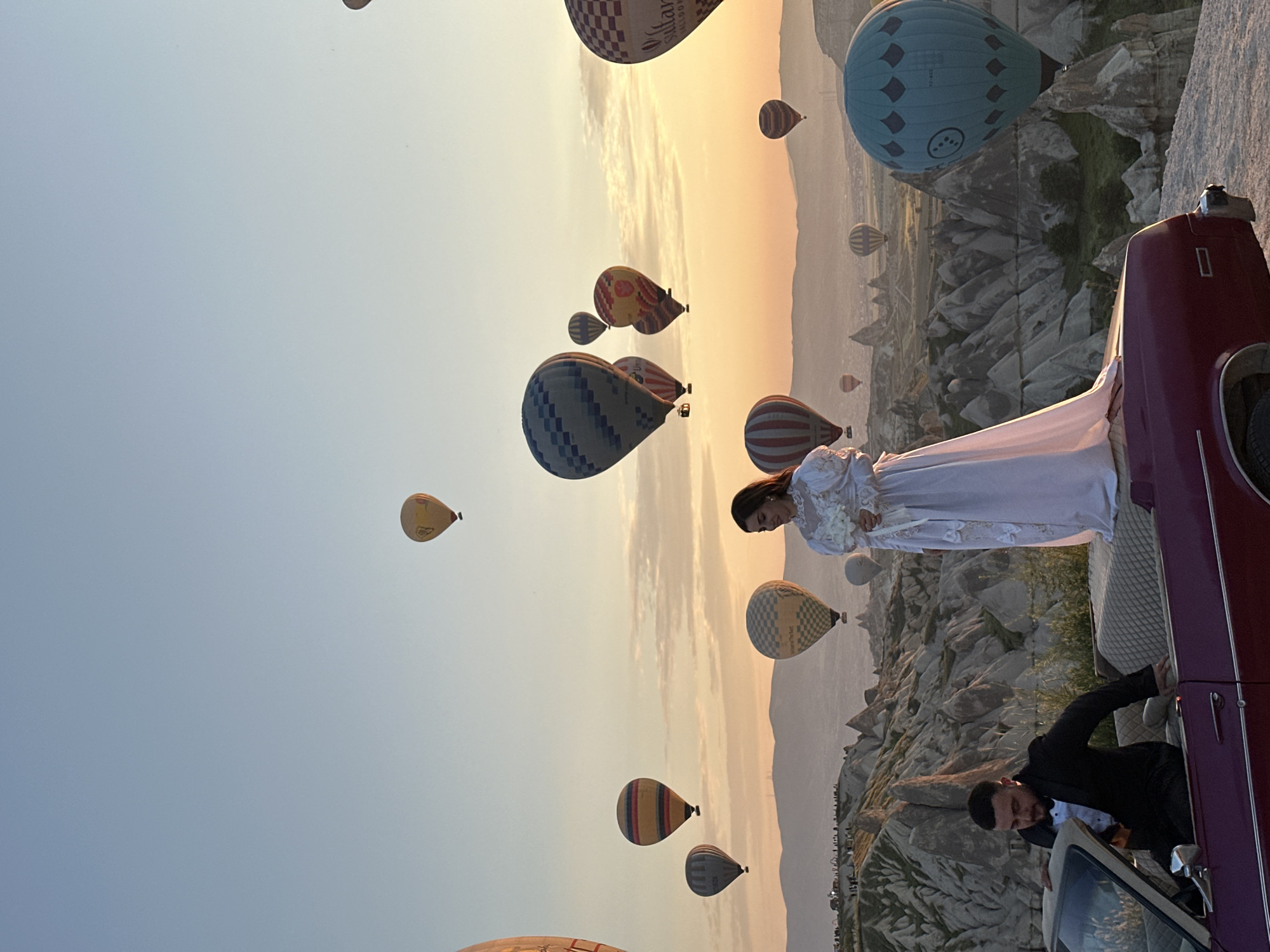 The image depicts a breathtaking sunrise or sunset scene in Göreme, Turkey, showcasing a couple posing with a vintage car amidst a sky filled with hot air balloons. The sky gradient shifts from a pale blue at the top to warm orange and yellow tones near the horizon, indicative of either early morning or late evening.
In the foreground, positioned on the right, is a vintage dark red/maroon car with chrome detailing. A woman, elegantly dressed in a white, possibly bridal, gown, stands gracefully on the rear of the vehicle, looking towards the left side of the frame. Her dark hair is neatly styled. Next to the car, a man in a black suit, white shirt, and bow tie leans out from the driver's side door, smiling slightly towards the viewer.
The midground and background reveal the distinctive rocky landscape of Cappadocia, characterized by its unique "fairy chimney" formations. Above this iconic scenery, numerous hot air balloons of various designs and colors ascend. Text visible on some balloons includes "SULTAN BALLOONS" and "ÜRGÜP" on a checkered balloon, "TC-BGH" on a blue balloon with diamond patterns, and "Royal Balloon" on a yellow balloon.
This scene appears to capture a special event, likely a wedding or engagement photoshoot, set against the spectacular backdrop of Cappadocia's hot air balloon display, commonly observed at sunrise. The overall atmosphere conveyed is romantic, celebratory, and serene.